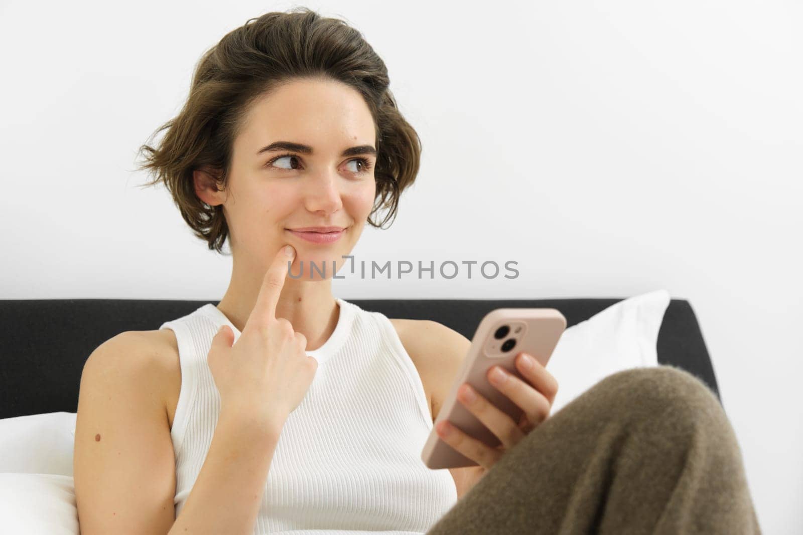 Close up portrait of smiling woman in bed, thinking, holding smartphone, looking aside with pleased smirking face, making decision, online shopping on mobile phone while spending time at home.