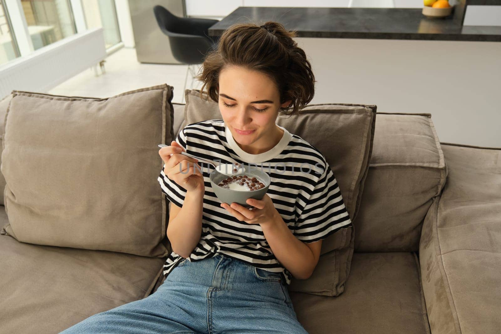 Portrait of young female model, student eating quick breakfast, holding bowl of granola or cereals with milk, sitting on sofa in living room by Benzoix