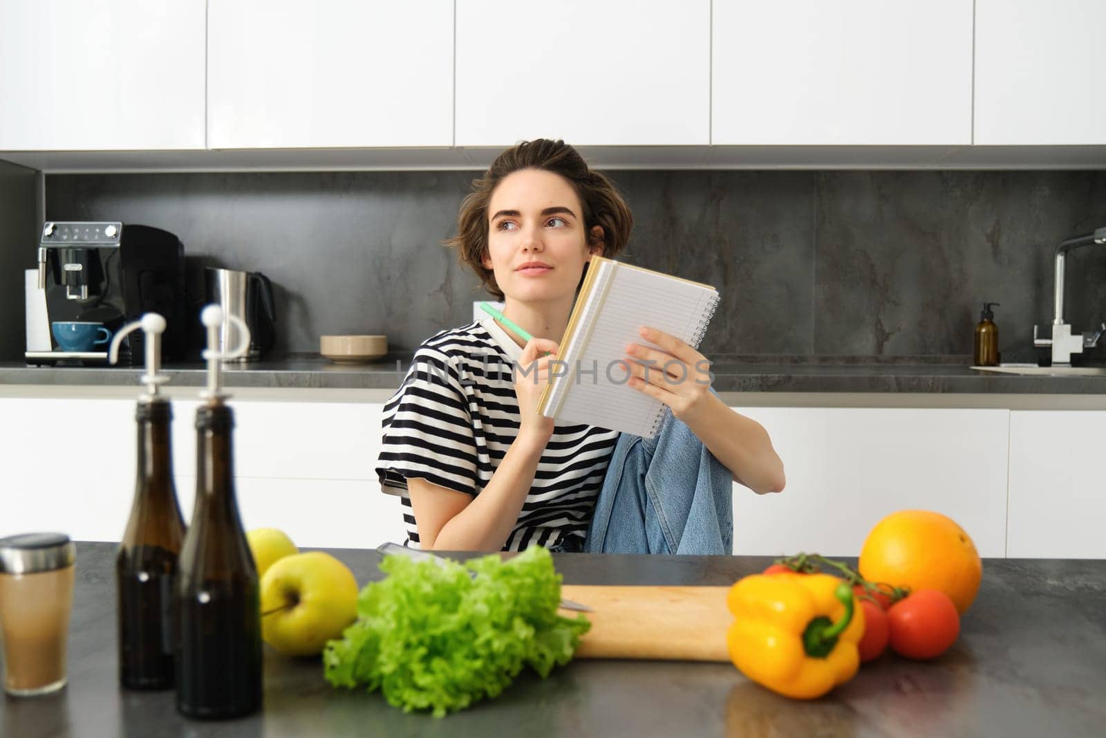 Portrait of young smiling woman in kitchen, holding notebook, making notes for recipe, writing grocery list, cooking salad, sitting near vegetables and chopping board.