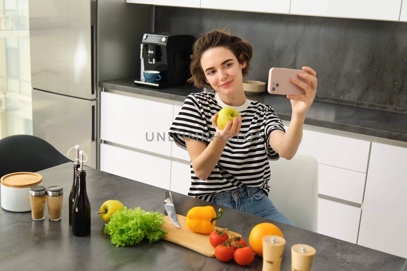 Portrait of beautiful young woman, food blogger recording video of her cooking in the kitchen, showing meal ingredients, preparing a meal on camera, using smartphone for live streaming.