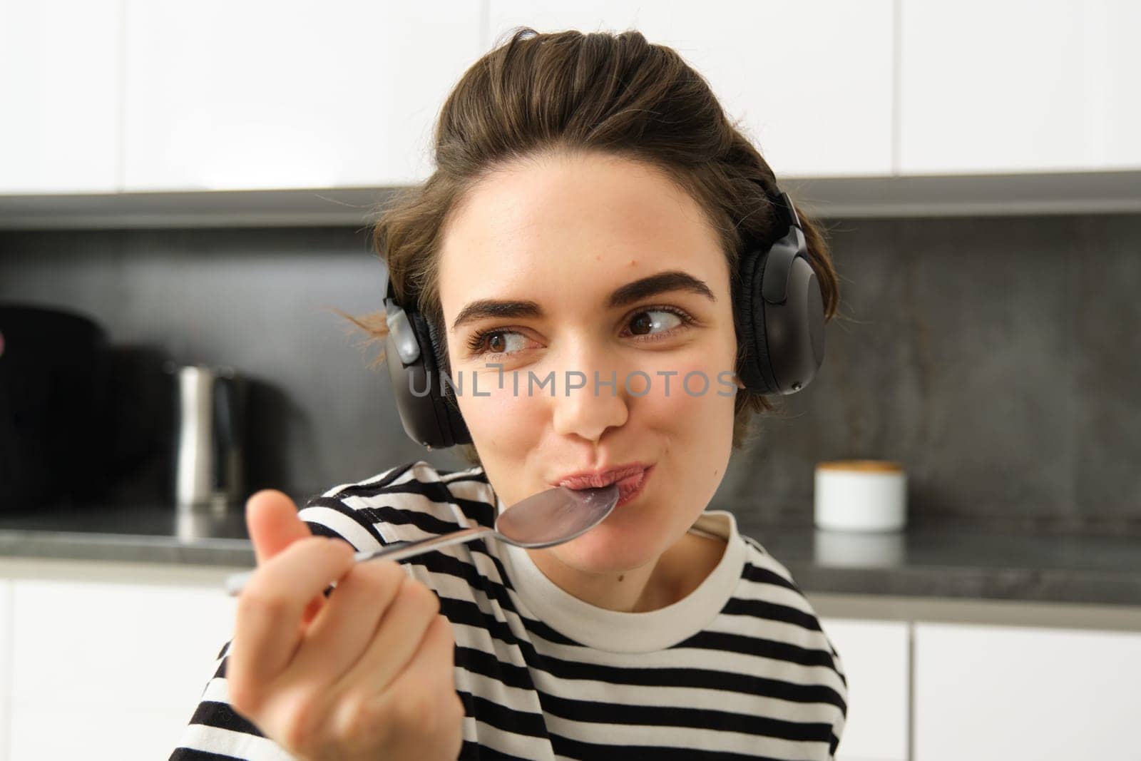 Close up portrait of young smiling woman in headphones, eating cereals with spoon and listening music, wearing earphones, sitting in the kitchen by Benzoix