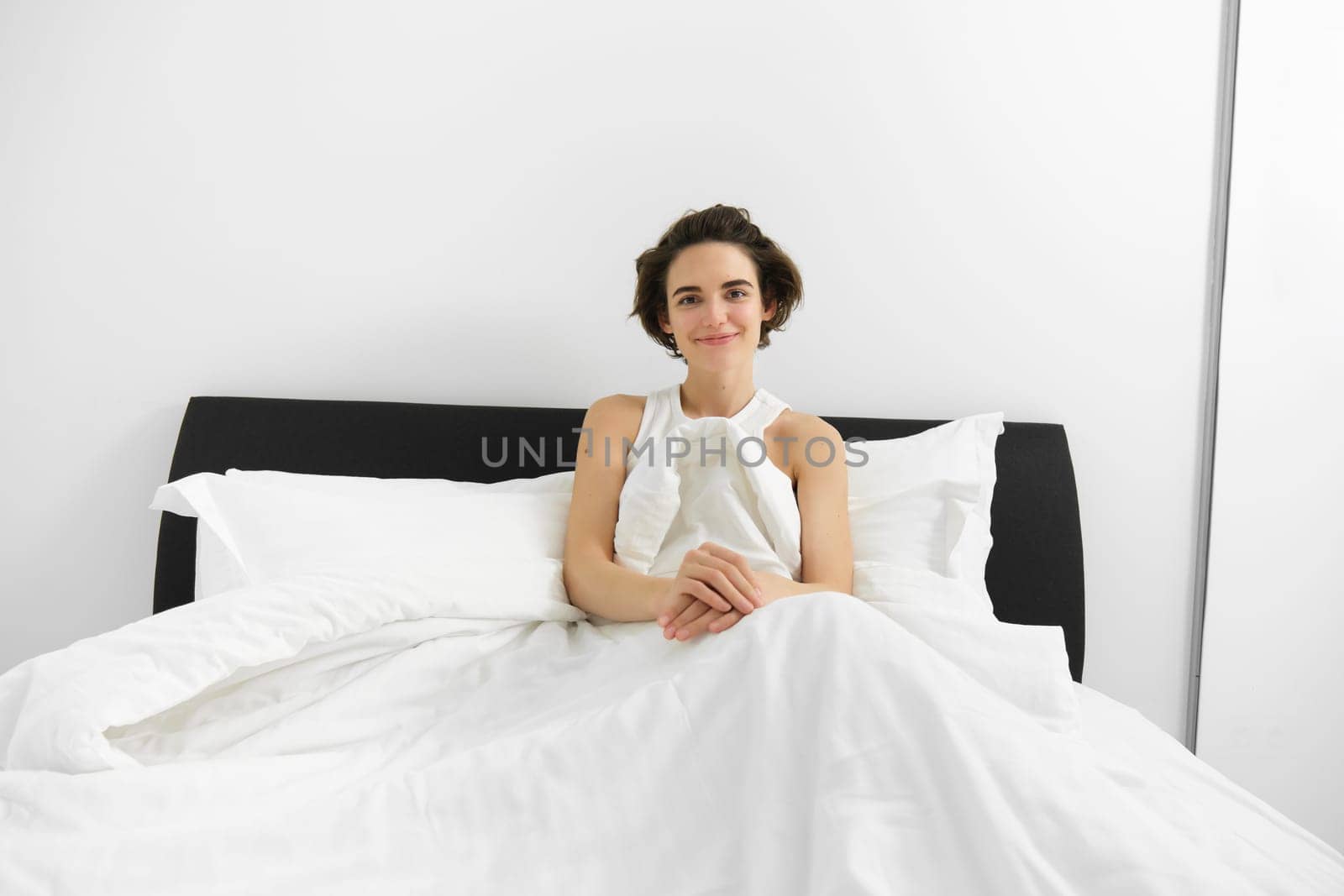 Image of smiling candid female model, lying in bed under white sheets, looking happy at camera, concept of morning, waking up early by Benzoix
