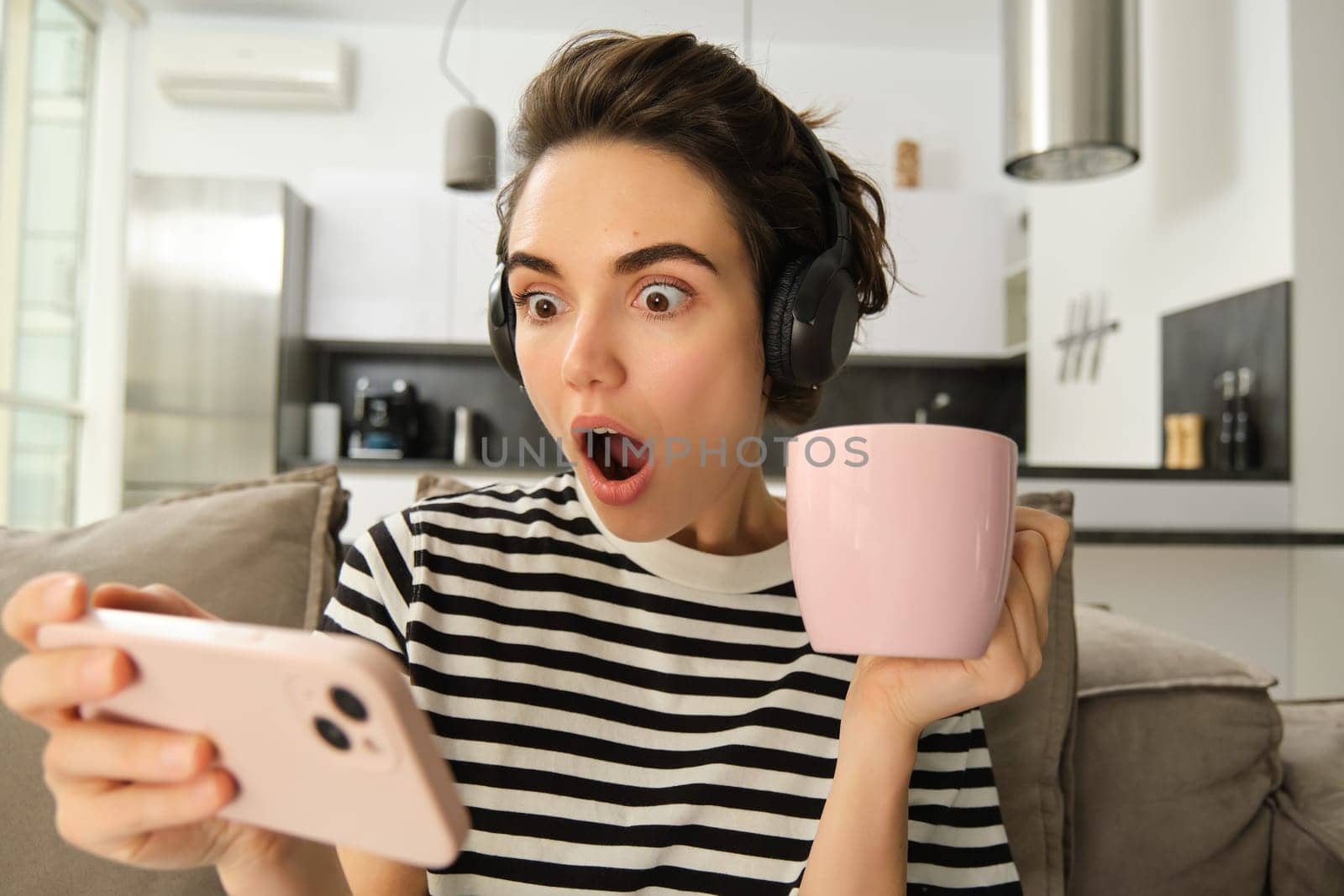 Portrait of woman binge watching her favourite tv show on smartphone, drinking tea and wearing headphones, gasping from shock, looking amazed at mobile screen.