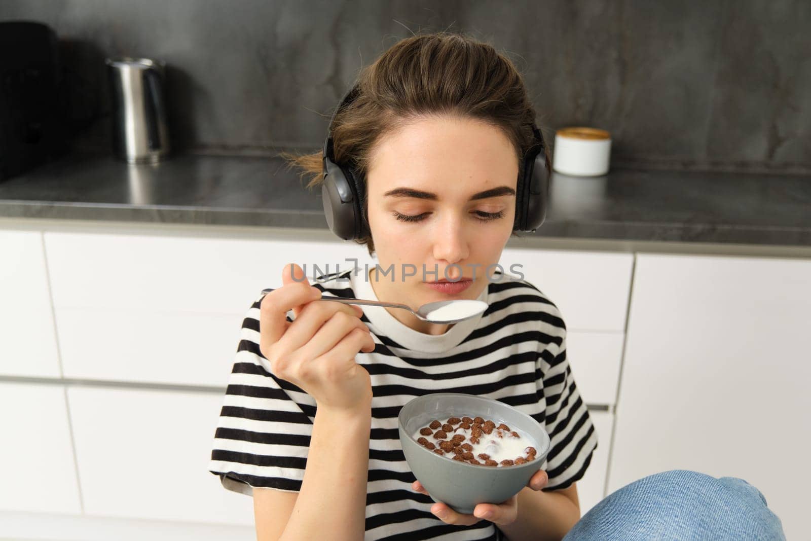 Image of stylish, modern woman, student eating cereals with milk for breakfast, holding bowl and spoon, listening music in wireless black headphones by Benzoix