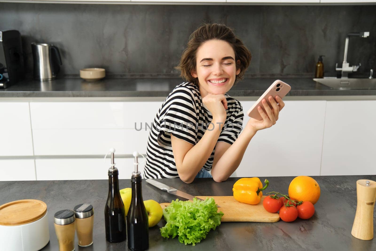 Happy smiling woman, sitting in the kitchen with smartphone, cooking salad, has chopping board, olive oil and vegetables on table.