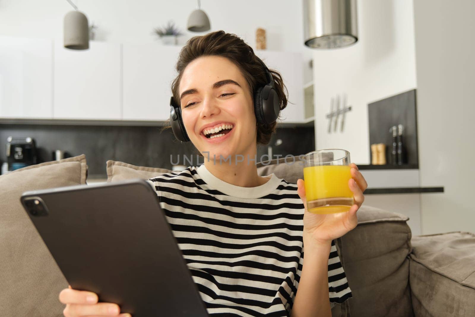 Portrait of woman in headphones, sitting on sofa with tablet and drinking orange juice, watching tv show on her gadget, using social media application, spending time at home.