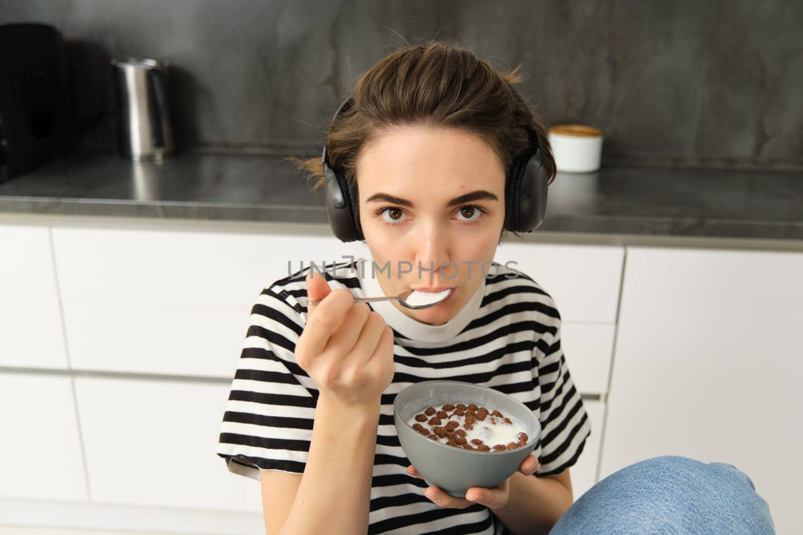 Beautiful woman, stylish girl eating cereals with milk, holding spoon near mouth and bowl in hands, having her breakfast in the morning, sitting in kitchen, listening music in headphones by Benzoix