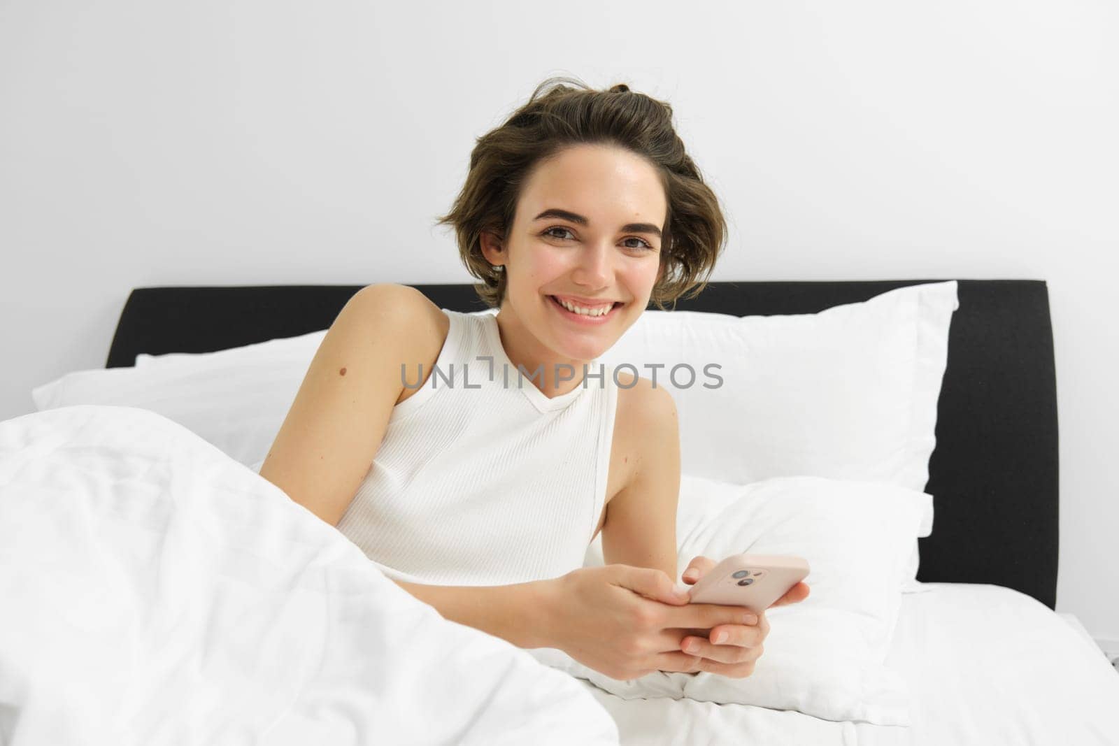 Image of woman checking her sleep app on mobile phone, lying in bed and smiling, using smartphone in bedroom by Benzoix