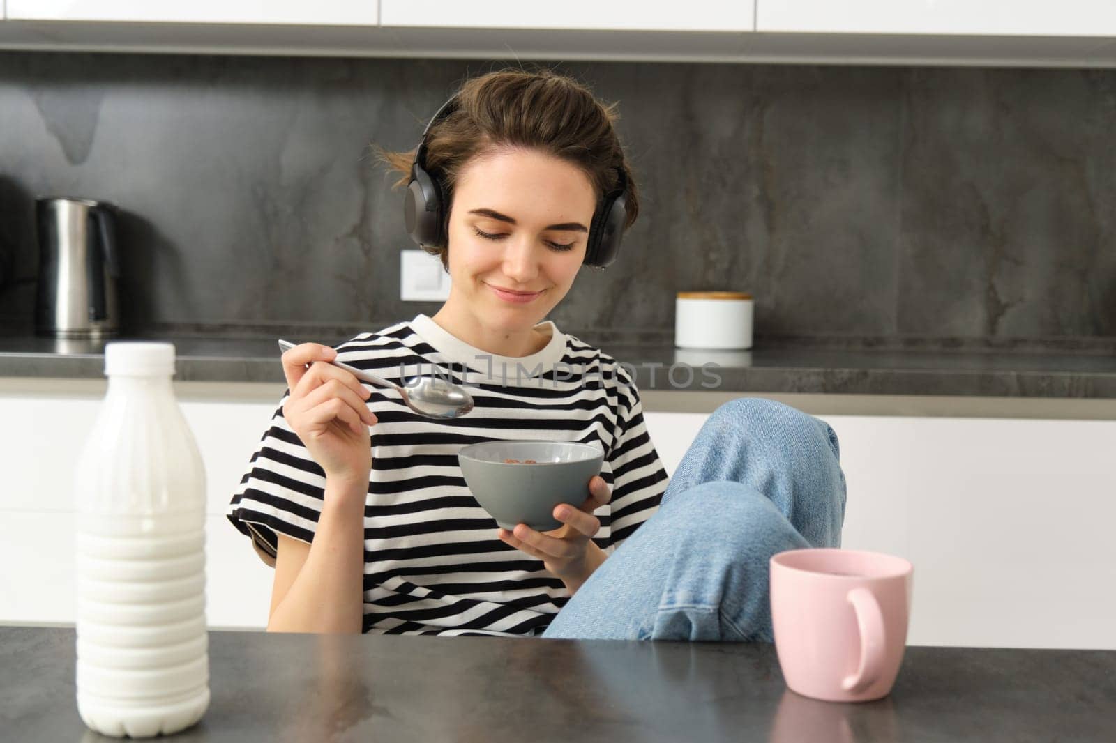 Portrait of smiling young candid woman, student eating morning cereals, having her breakfast, listening music in wireless headphones, sitting in the kitchen by Benzoix