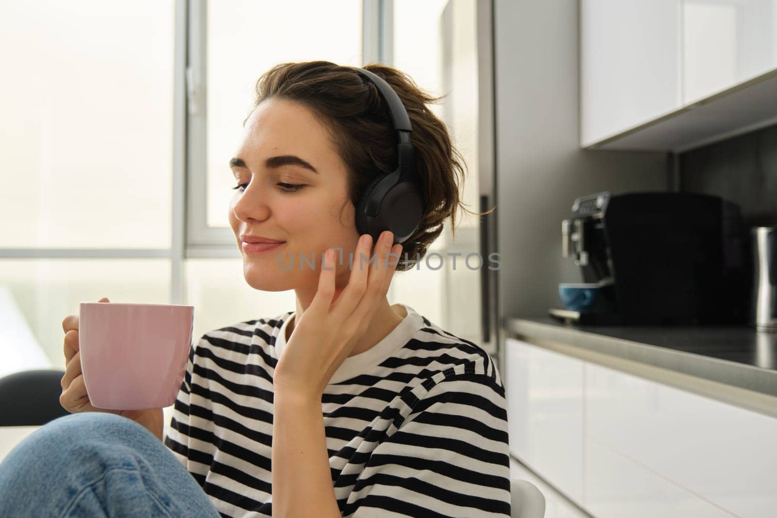 Close up portrait of smiling brunette woman, student in headphones, listens music, drinks cup of tea in morning, sits in kitchen and enjoys her daily routine by Benzoix