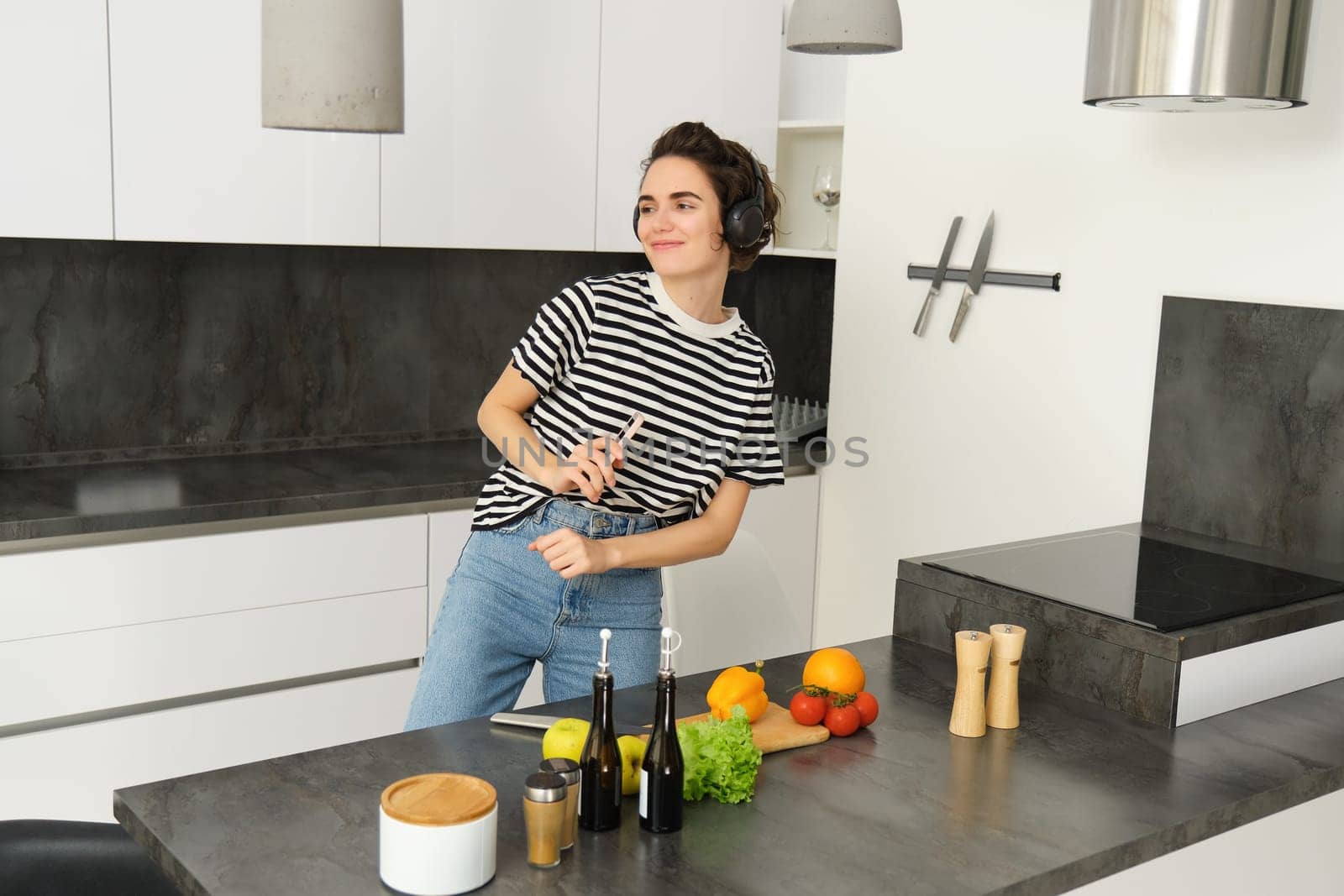 Happy woman dancing and cooking in the kitchen, listening music in earphones, making a meal, preparing salad, chopping vegetables on a counter by Benzoix