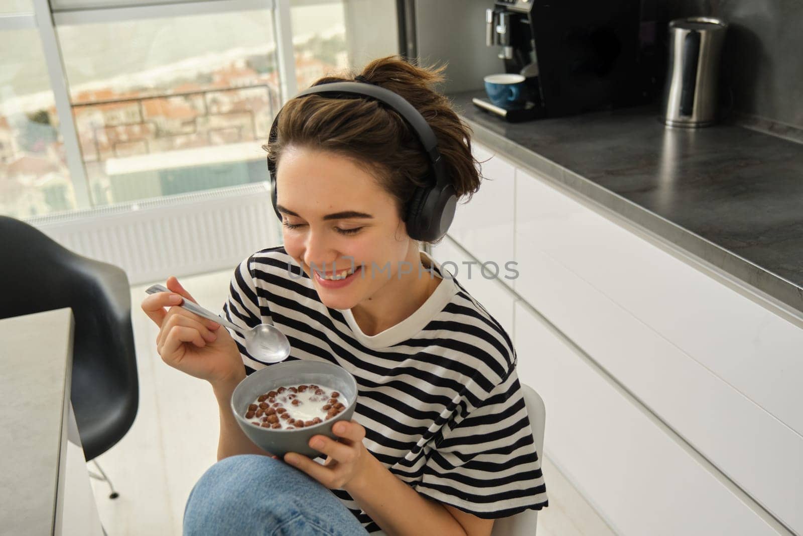 Close up portrait of smiling, beautiful young woman in headphones, eating cereals for breakfast and listening music or e-book, holding spoon with bowl by Benzoix