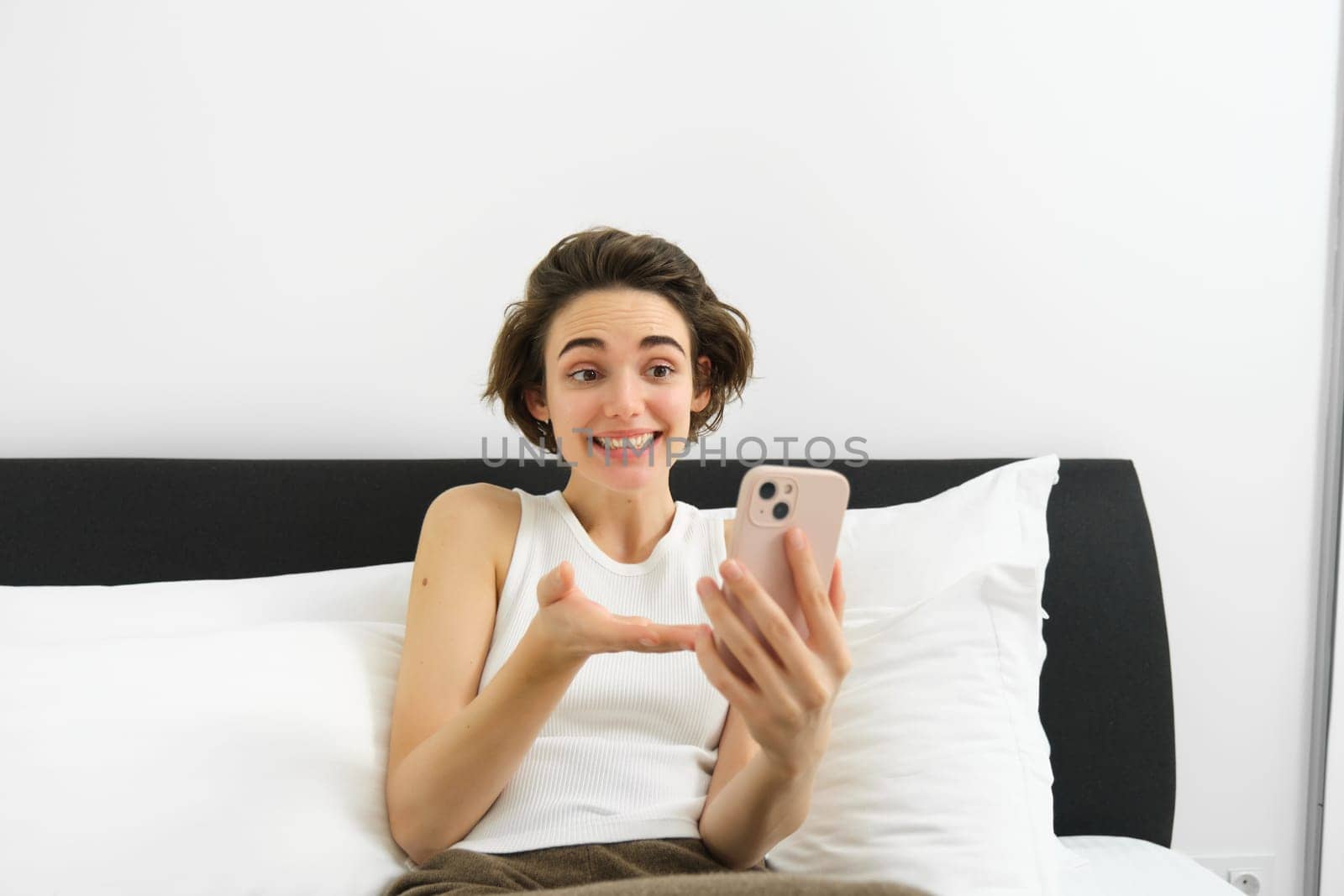 Cheerful smiling woman resting in bed, connects to online video call while lying in bedroom, talking to friend via smartphone app.