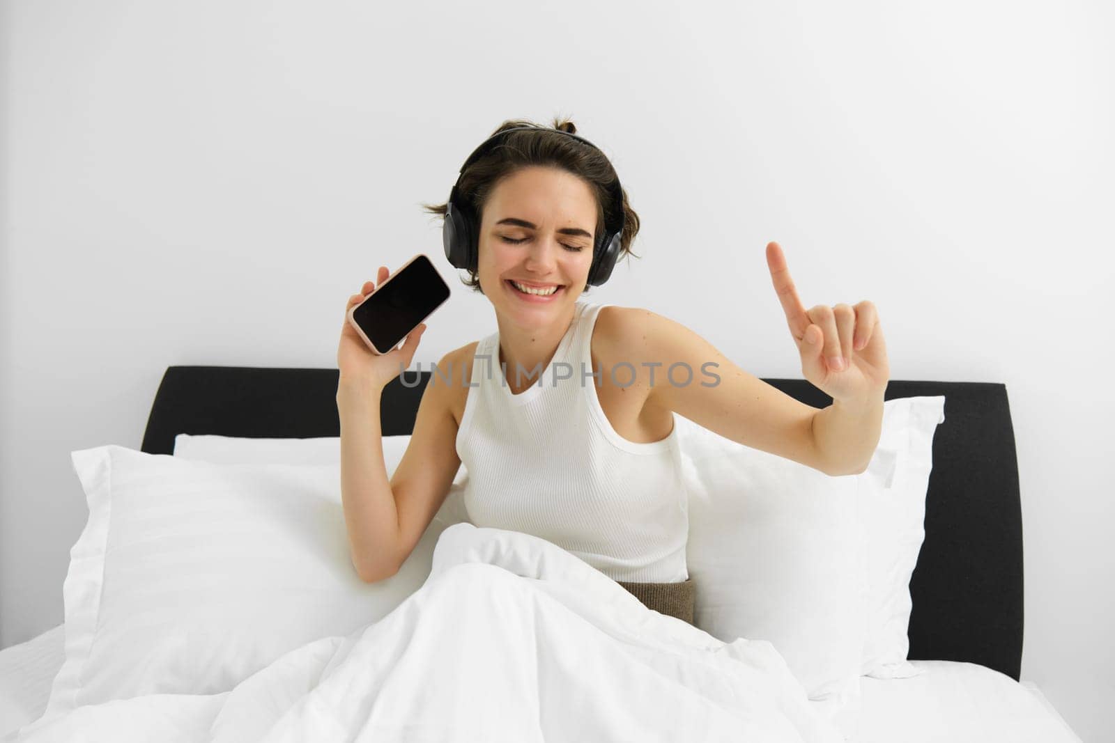 Image of beautiful smiling woman, dancing on bed, waking up and listening to music in wireless headphones, energizing, happy morning by Benzoix
