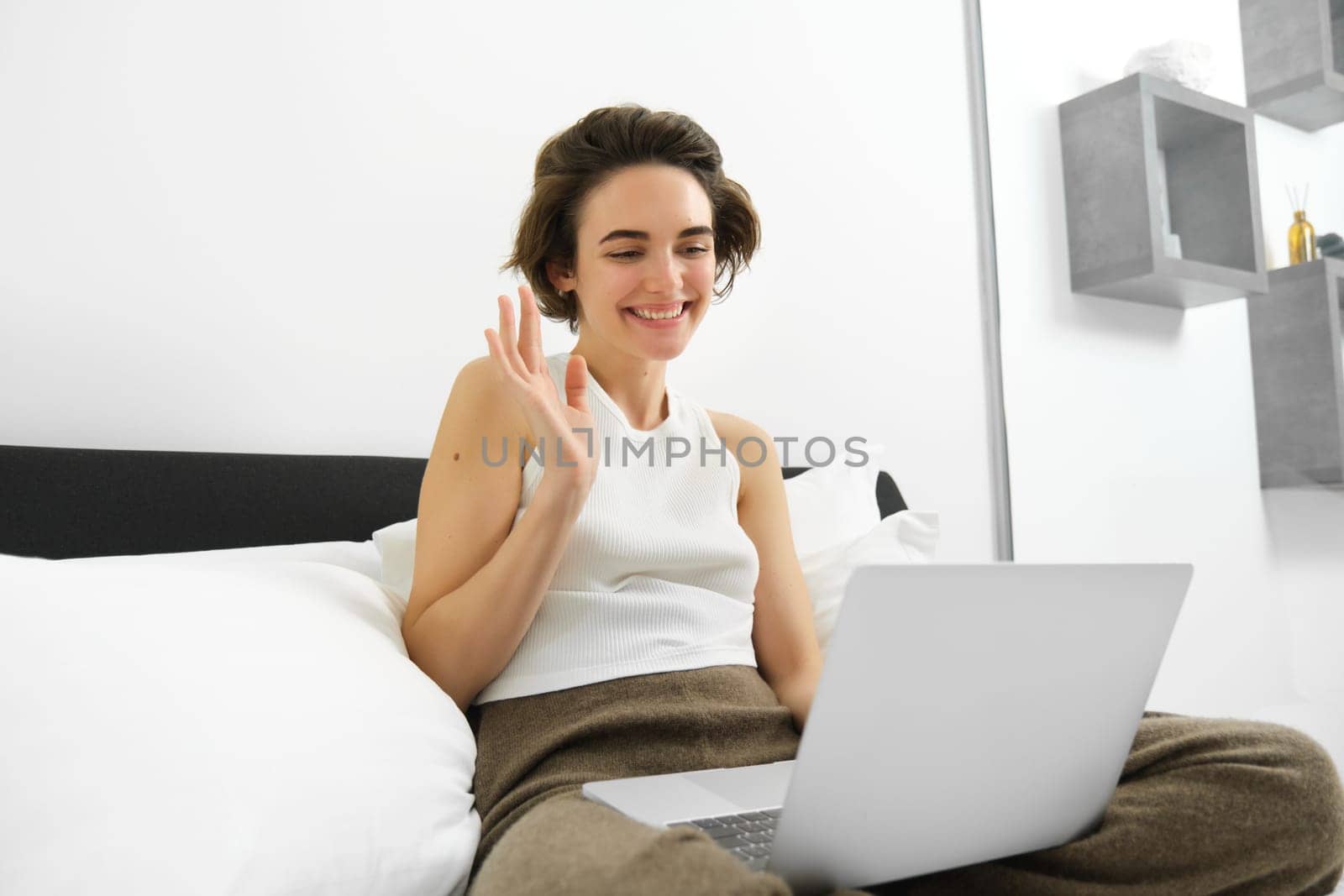 Image of young woman working from home, connects to online video chat, team meeting, waving hand at laptop, sitting on bed, studying, e-learning from home.