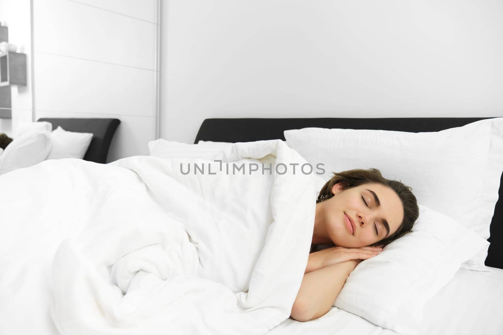 Image of beautiful young woman sleeping on comfortable bed, covered with soft white linen sheets, eyes closed, face is calm and unbothered.