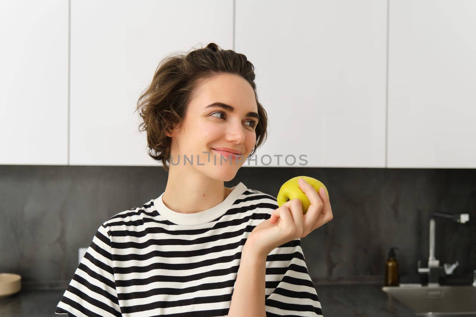 Portrait of beautiful, smiling young woman, holding an apple, eating fruit in the kitchen, looking happy. Concept of healthy diet and lifestyle.