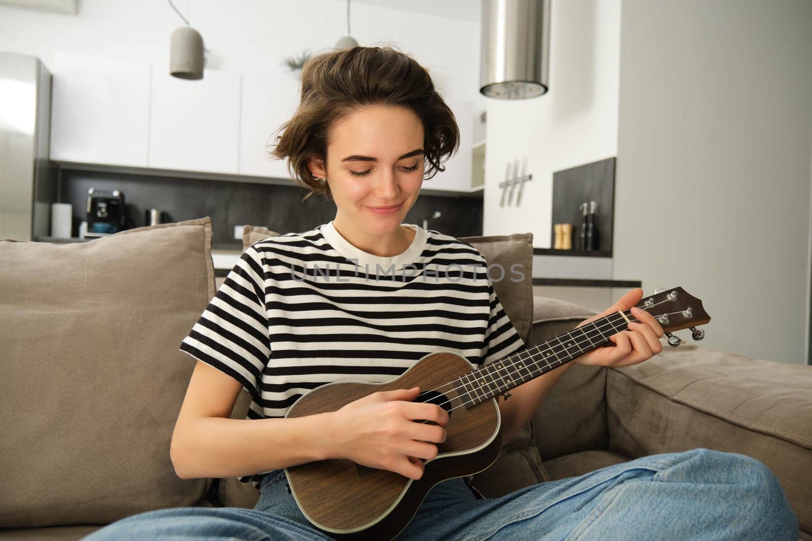 Portrait of young modern woman, student playing ukulele at home, sitting with small guitar, singing and feeling happy, sitting on sofa. Lifestyle and music concept