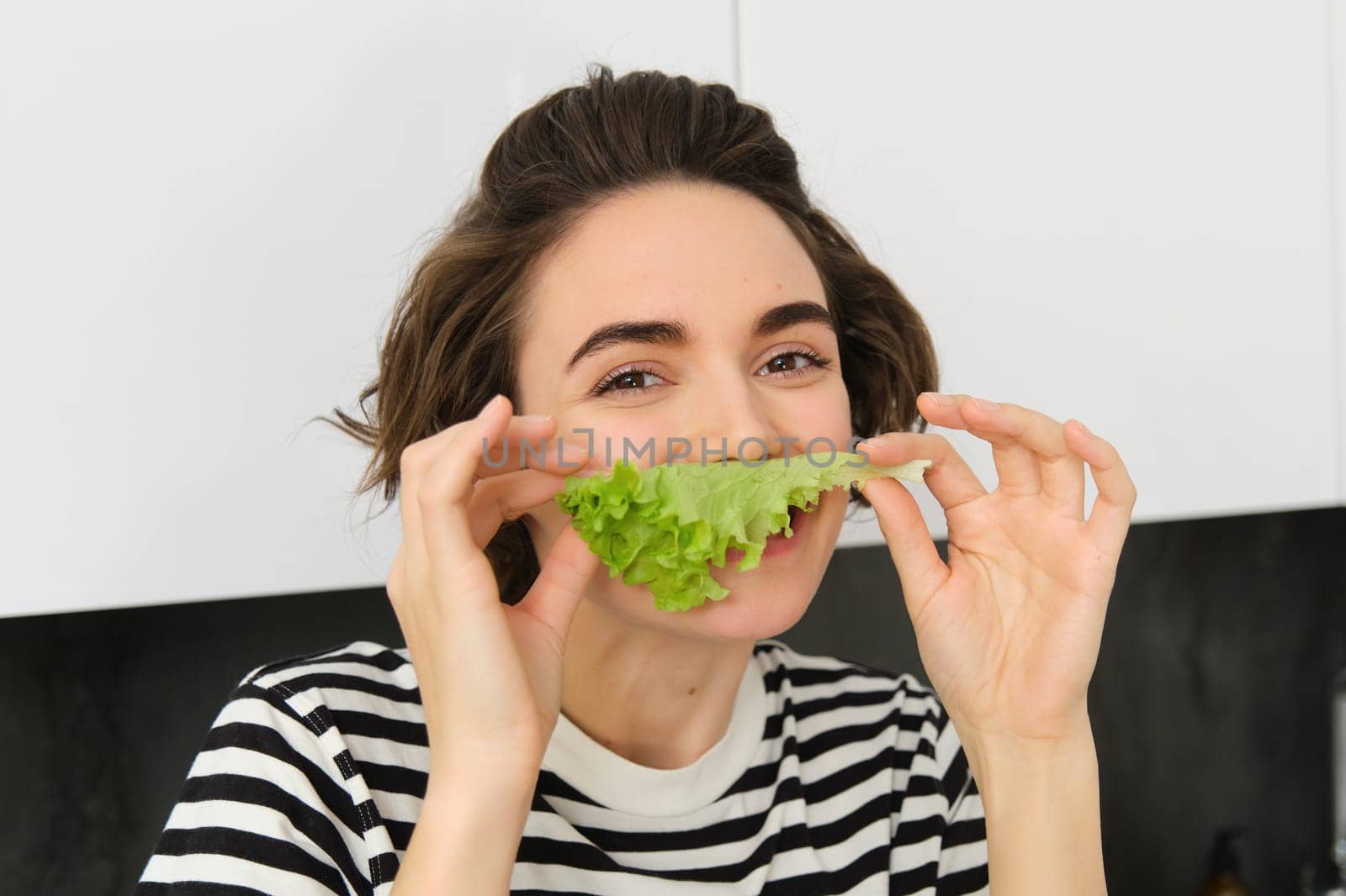 Close up portrait of young woman, vegetarian girl, likes eating vegetables, posing with lettuce leaf and smiling, posing in the kitchen. Concept of healthy food and diet by Benzoix
