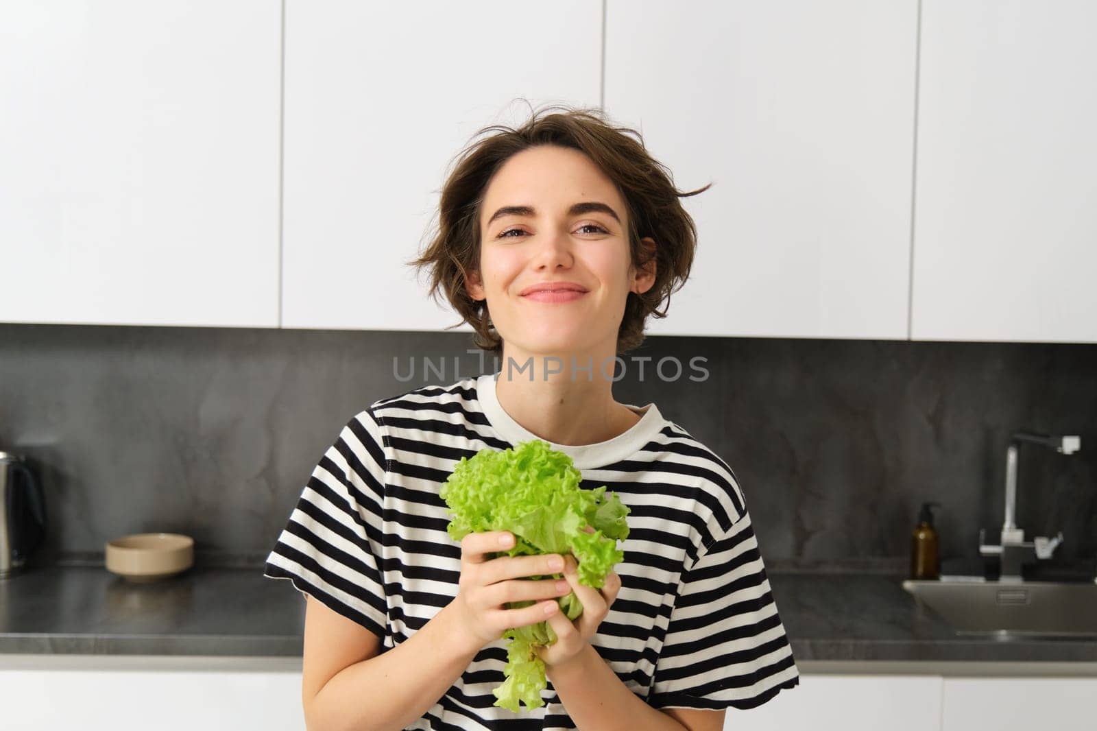 Portrait of happy and healthy young woman, following her diet, posing with lettuce leaf and smiling, cooking in the kitchen, vegetarian loves her vegetables.