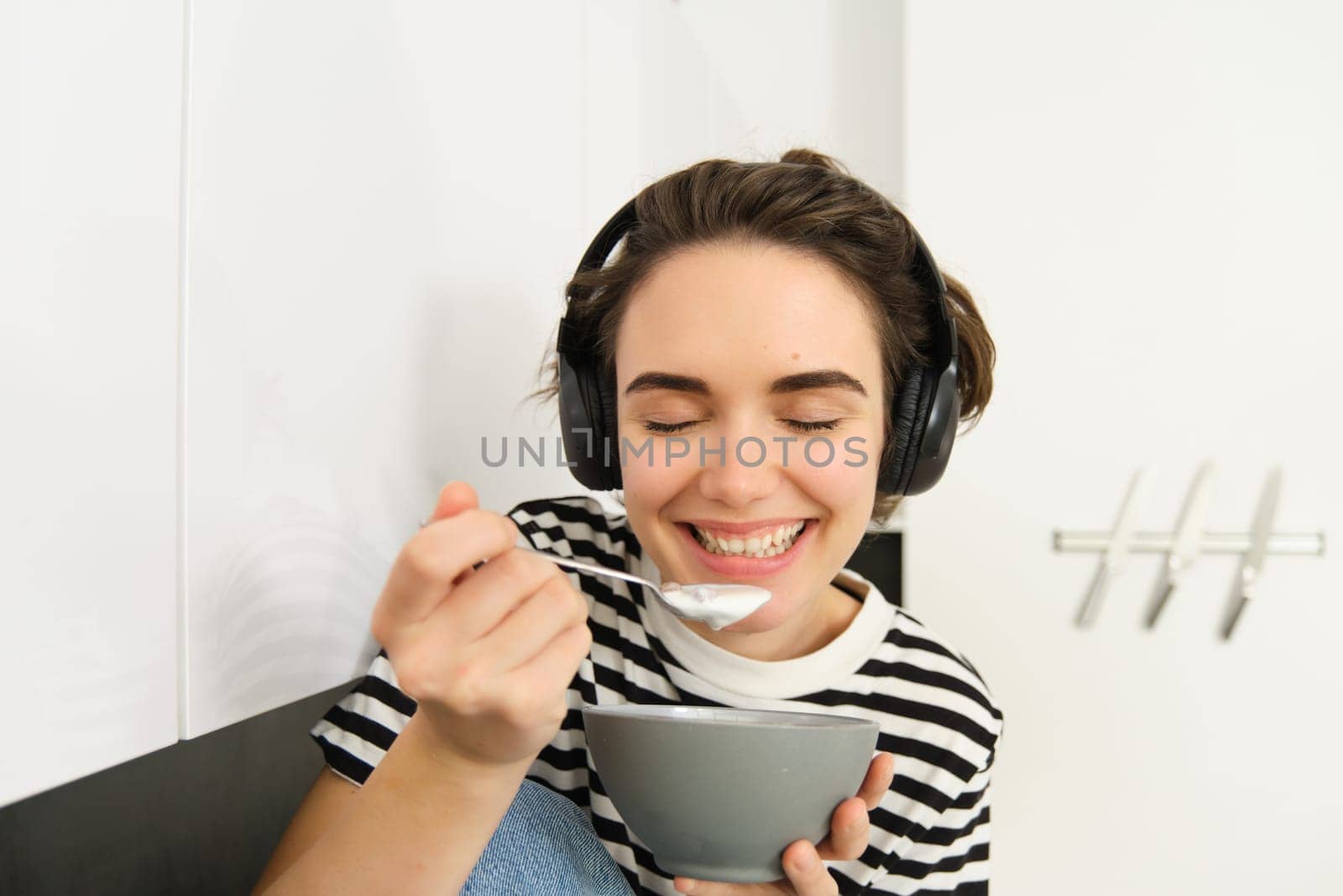 Happy smiling woman eats cereals with pleasure, holds bowl and spoon, listens music in headphones, sits on kitchen counter. Lifestyle concept