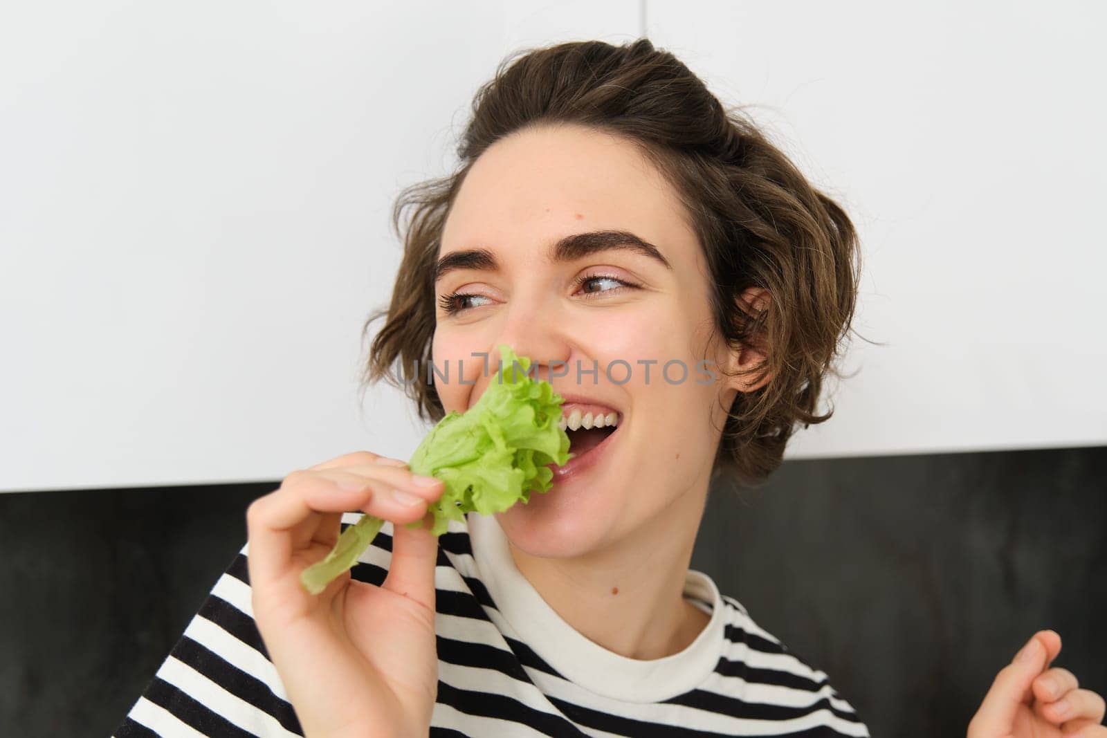 Portrait of carefree vegetarian girl, eating vegies, bite lettuce leaf with happy smiling face, having healthy snack, likes vegetables, stands in the kitchen.
