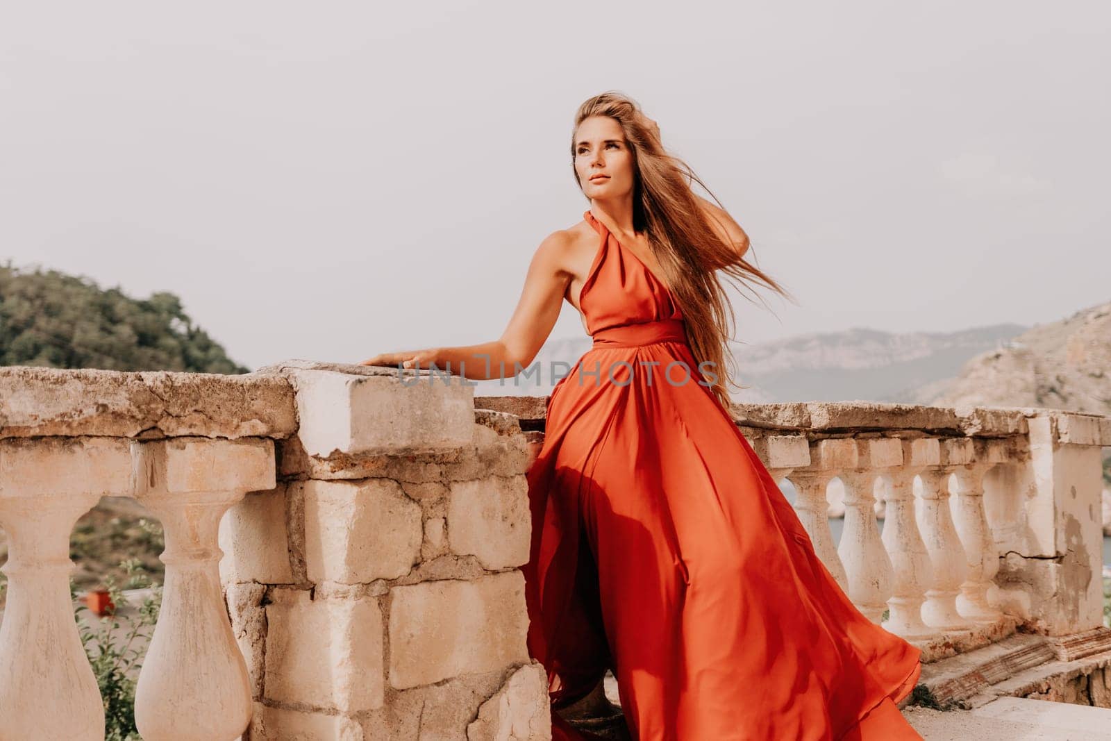 Woman travel portrait. Happy woman with long hair looking at camera and smiling. Close up portrait cute woman in a res long dress posing on backdrop of old travel city near sea by panophotograph