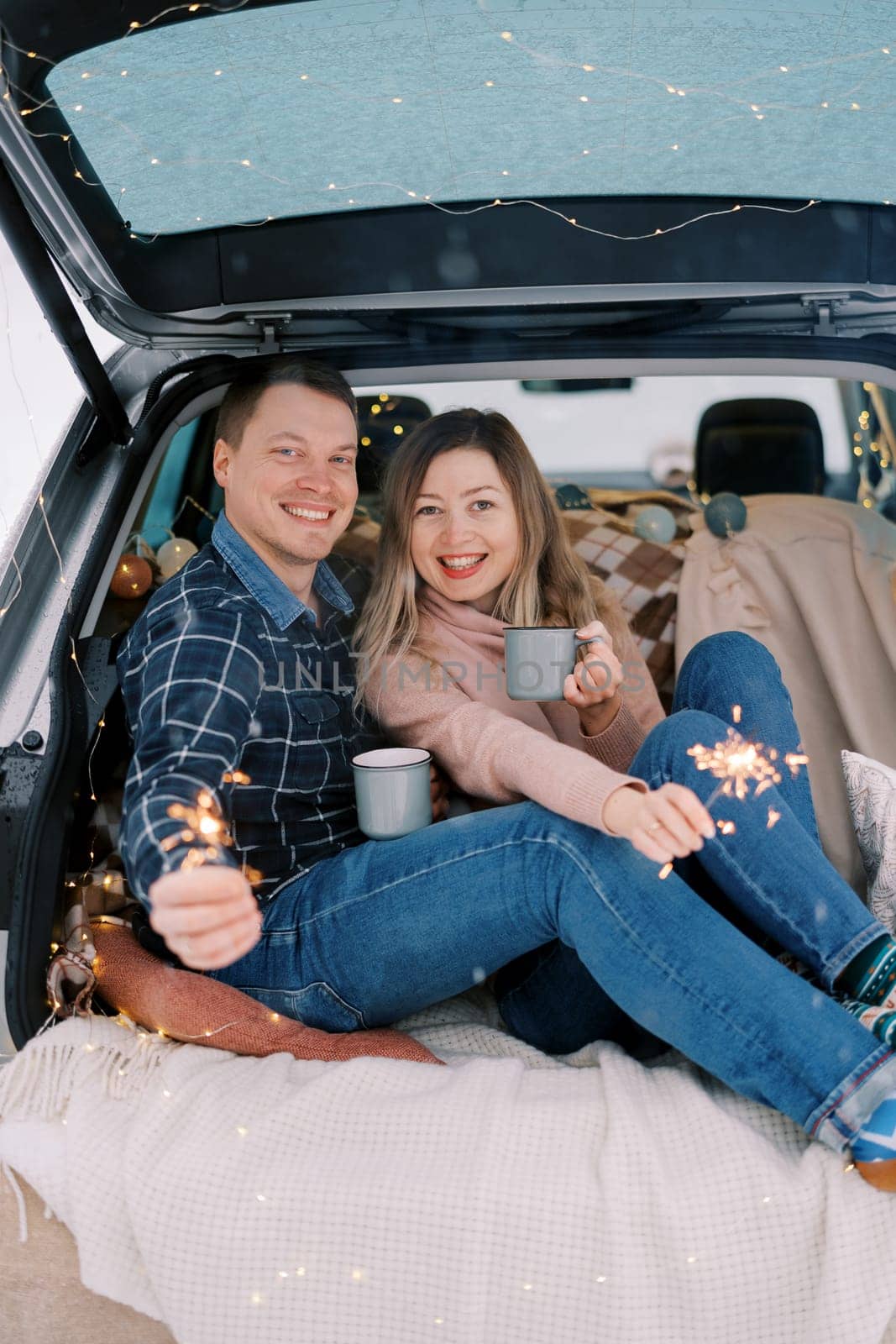 Smiling couple with mugs of coffee and sparklers in their hands are sitting on bedspreads in the trunk of a car by Nadtochiy