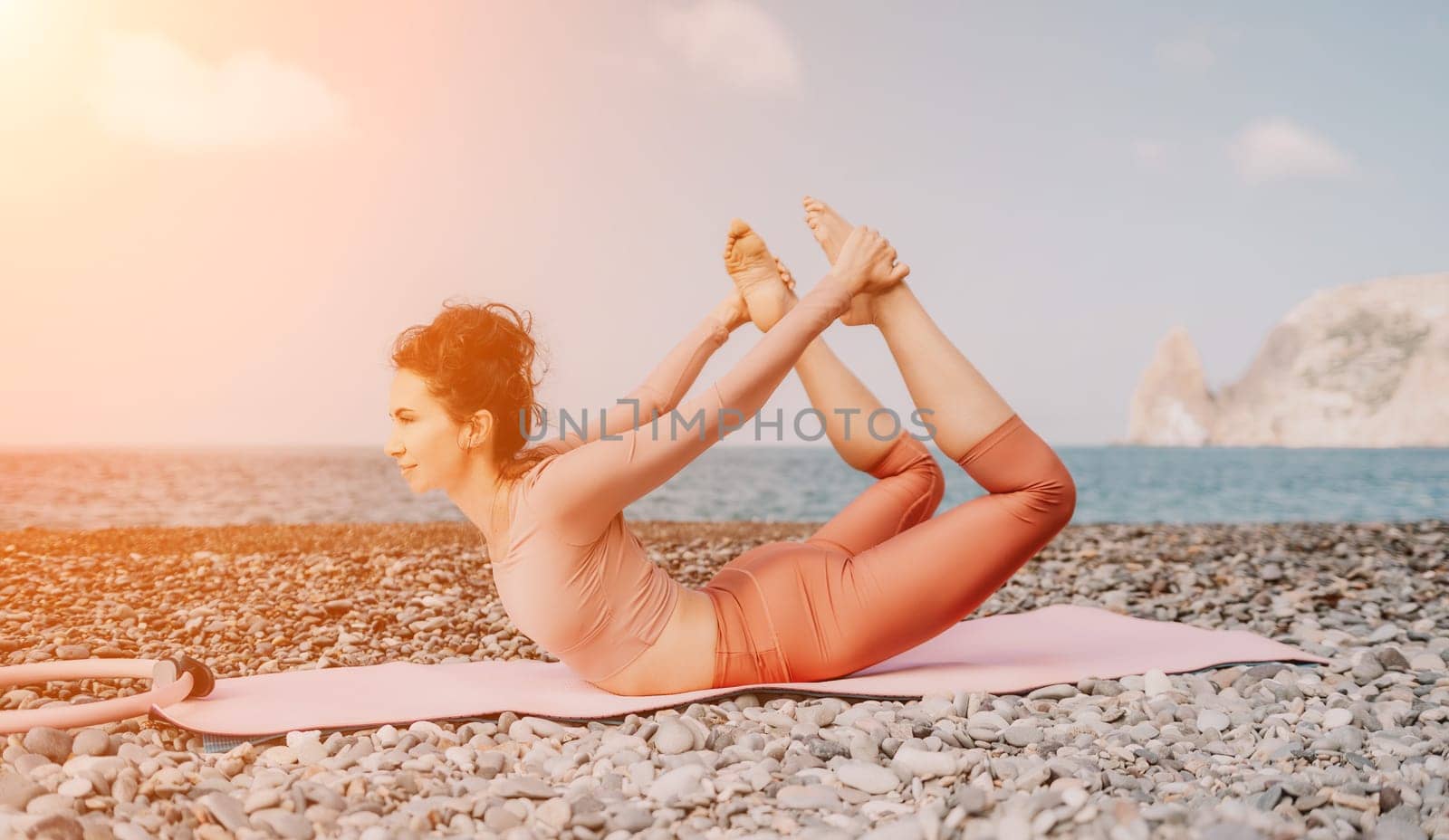 Middle aged well looking woman with black hair doing Pilates with the ring on the yoga mat near the sea on the pebble beach. Female fitness yoga concept. Healthy lifestyle, harmony and meditation.