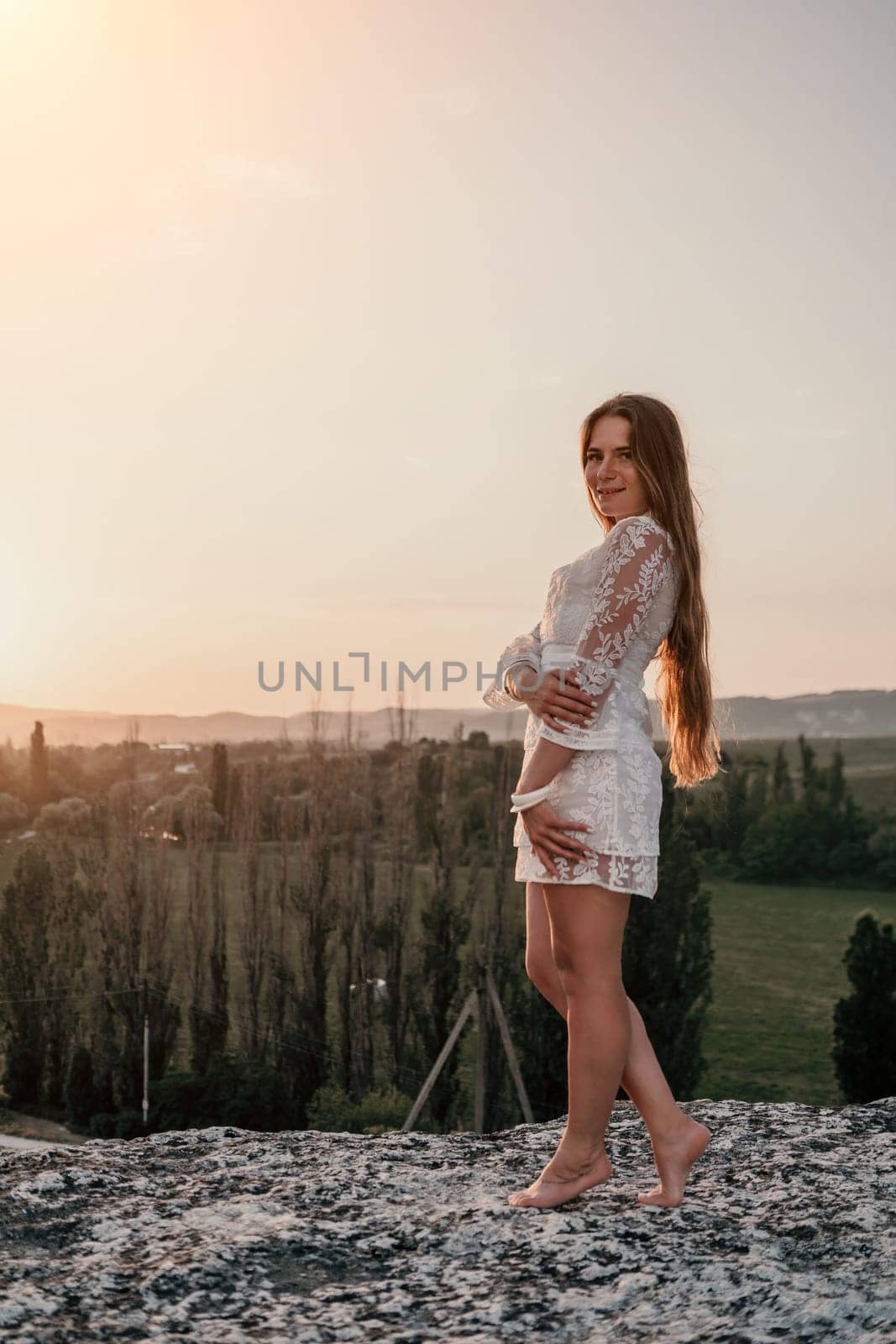 Romantic beautiful bride in white dress posing with sea and mountains in background. Stylish bride standing back on beautiful landscape of sea and mountains on sunset