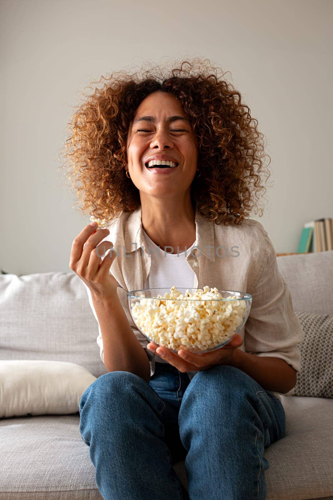 Vertical portrait of young multiracial hispanic woman laughing watching tv at home sitting on the sofa eating popcorn. Leisure activity and lifestyle concept.