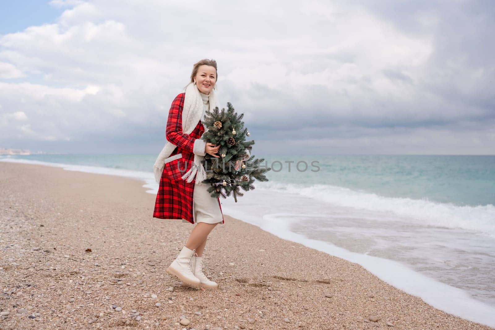 Blond woman holding Christmas tree by the sea. Christmas portrait of a happy woman walking along the beach and holding a Christmas tree in her hands. Dressed in a red coat, white dress