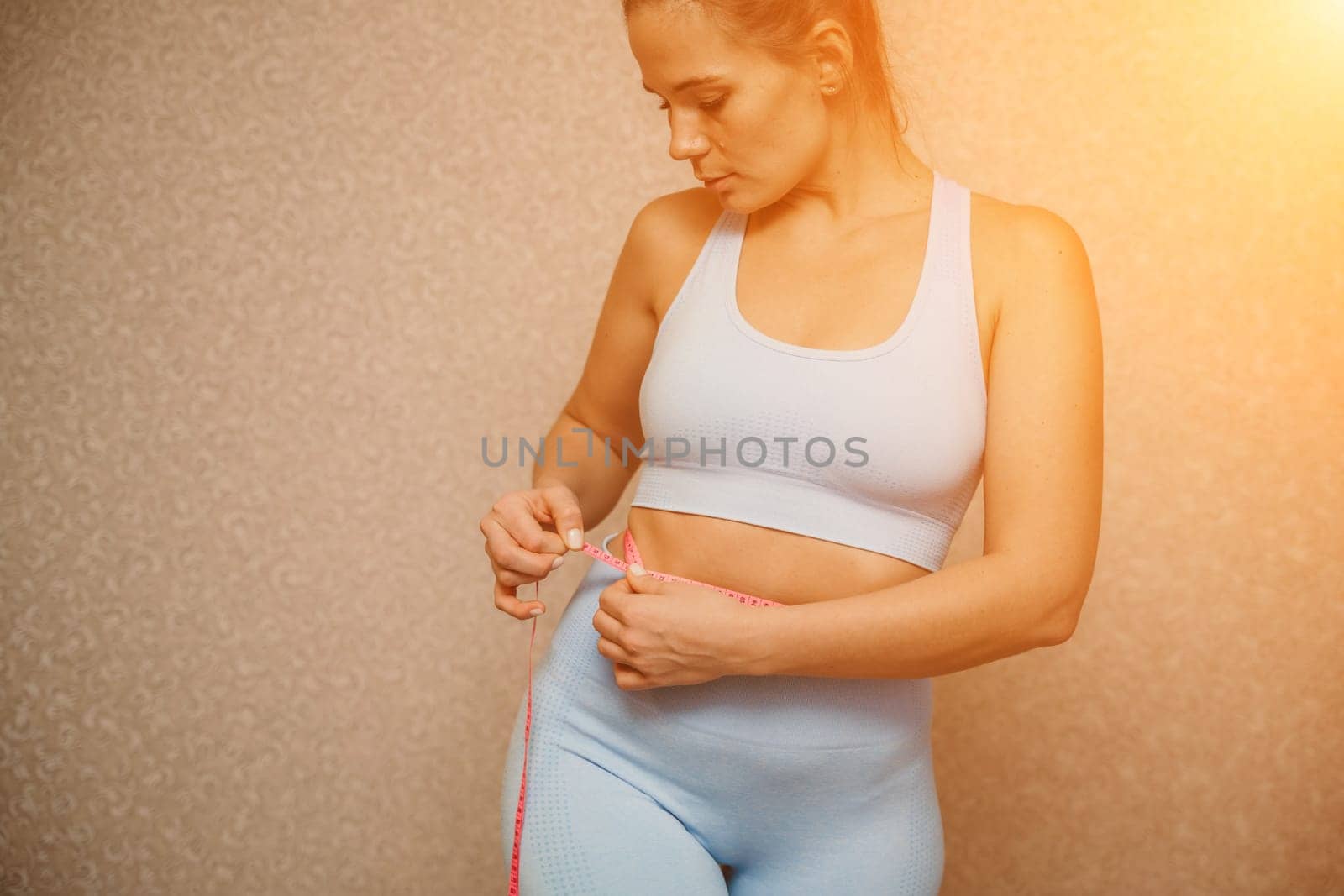 Cropped view of slim woman measuring waist with tape measure at home, close up. European woman checking the result of diet for weight loss or liposuction indoors.