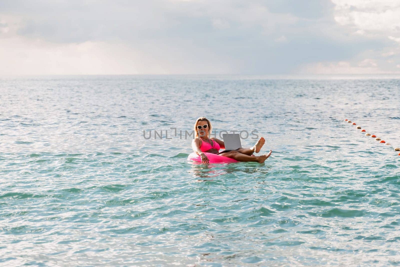 Woman works on laptop in sea. Freelancer, blond woman in sunglases floating on an inflatable big pink donut with a laptop in the sea at sunset. People summer vacation rest lifestyle concept