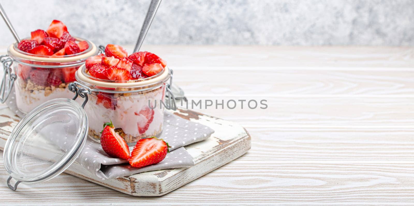 Parfait with Fresh Strawberries, Yoghurt and Crunchy Granola in Transparent Glass Mason Jars on White Rustic Wooden Background Angle View, Healthy Breakfast or Light Summer Dessert, Space for Text by its_al_dente