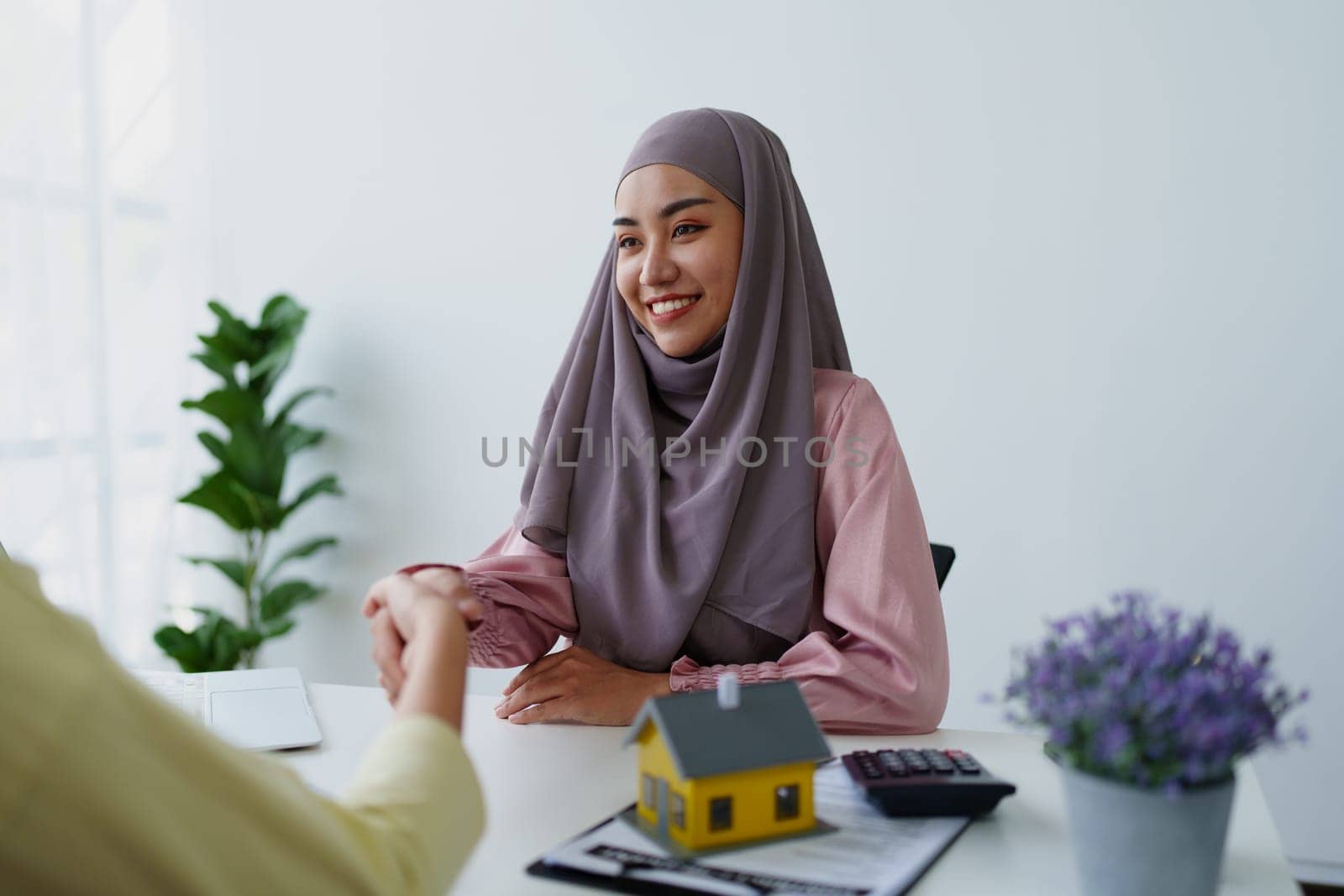 A female Muslim bank employee, holding hands, negotiates a residential loan with a customer. by Manastrong