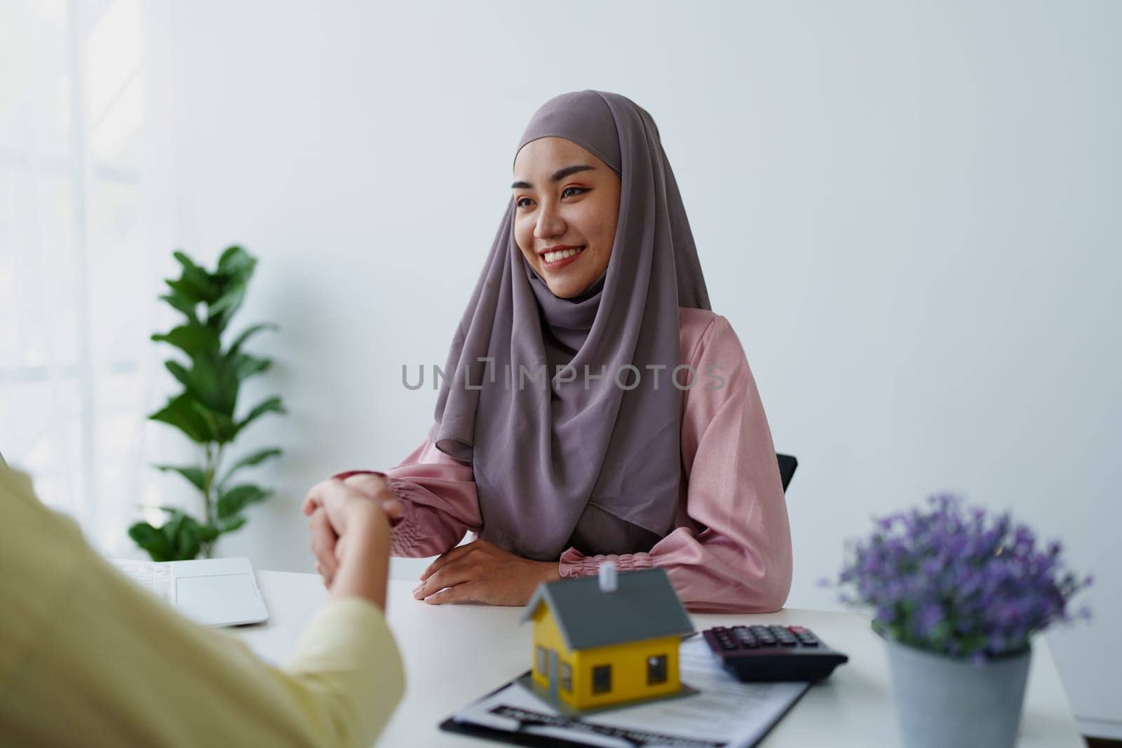 A female Muslim bank employee, holding hands, negotiates a residential loan with a customer