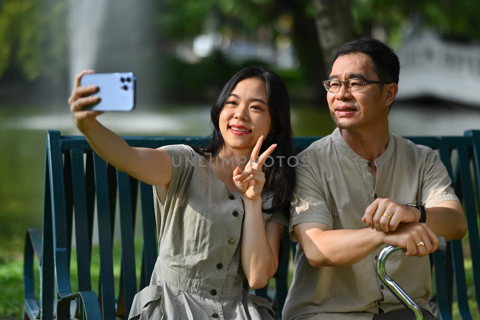 Happy smiling young woman with her father sitting on a wooden bench at public park making selfie on smartphone by prathanchorruangsak