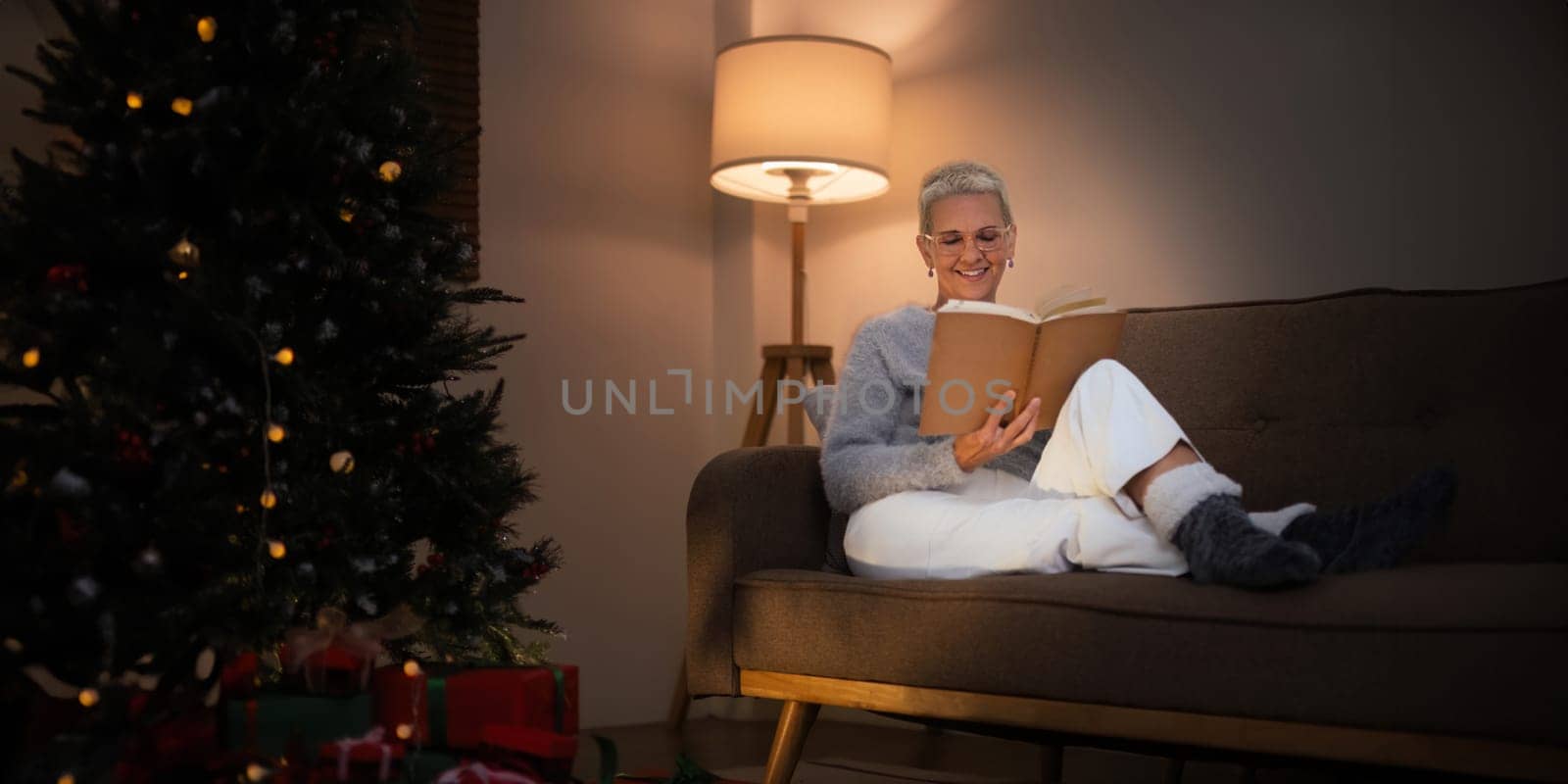 Smiling elderly woman with a cup of tea and a book relax in side of a decorated Christmas tree in living room.