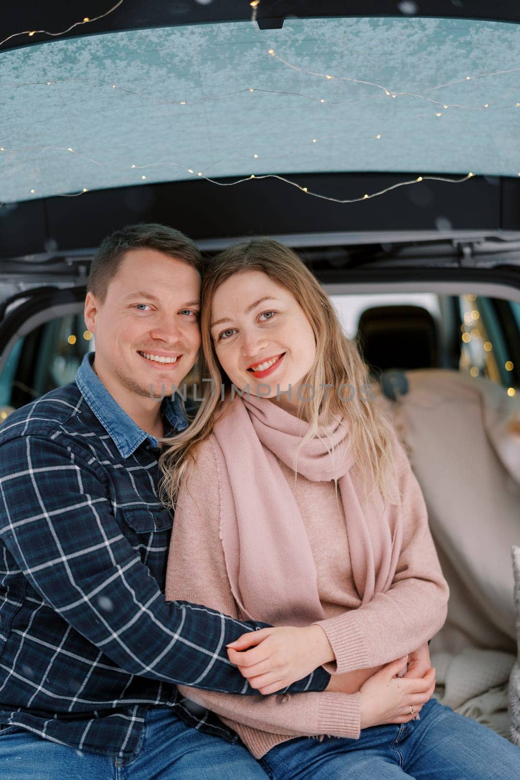 Hugging smiling couple sitting in car trunk on bedspreads by Nadtochiy