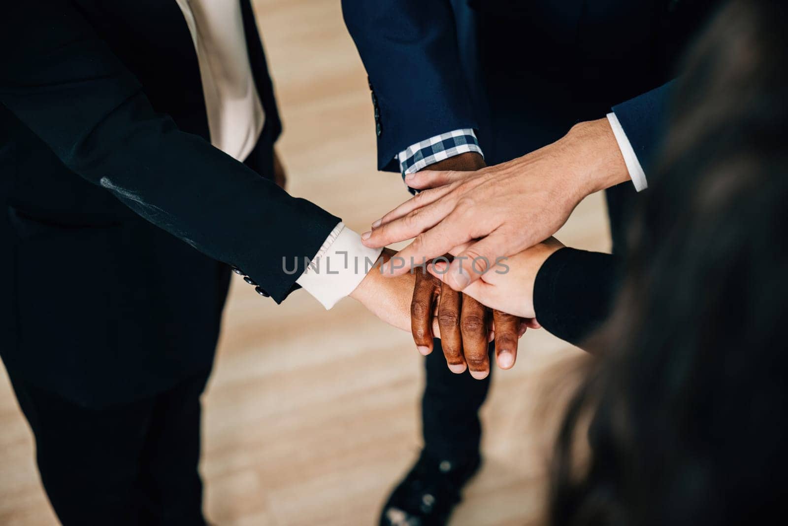 Business professionals gather in a modern office, their hands stacked in a circle as a symbol of achievement and unity. The scene exemplifies cooperation, communication, and a diverse workforce. by Sorapop