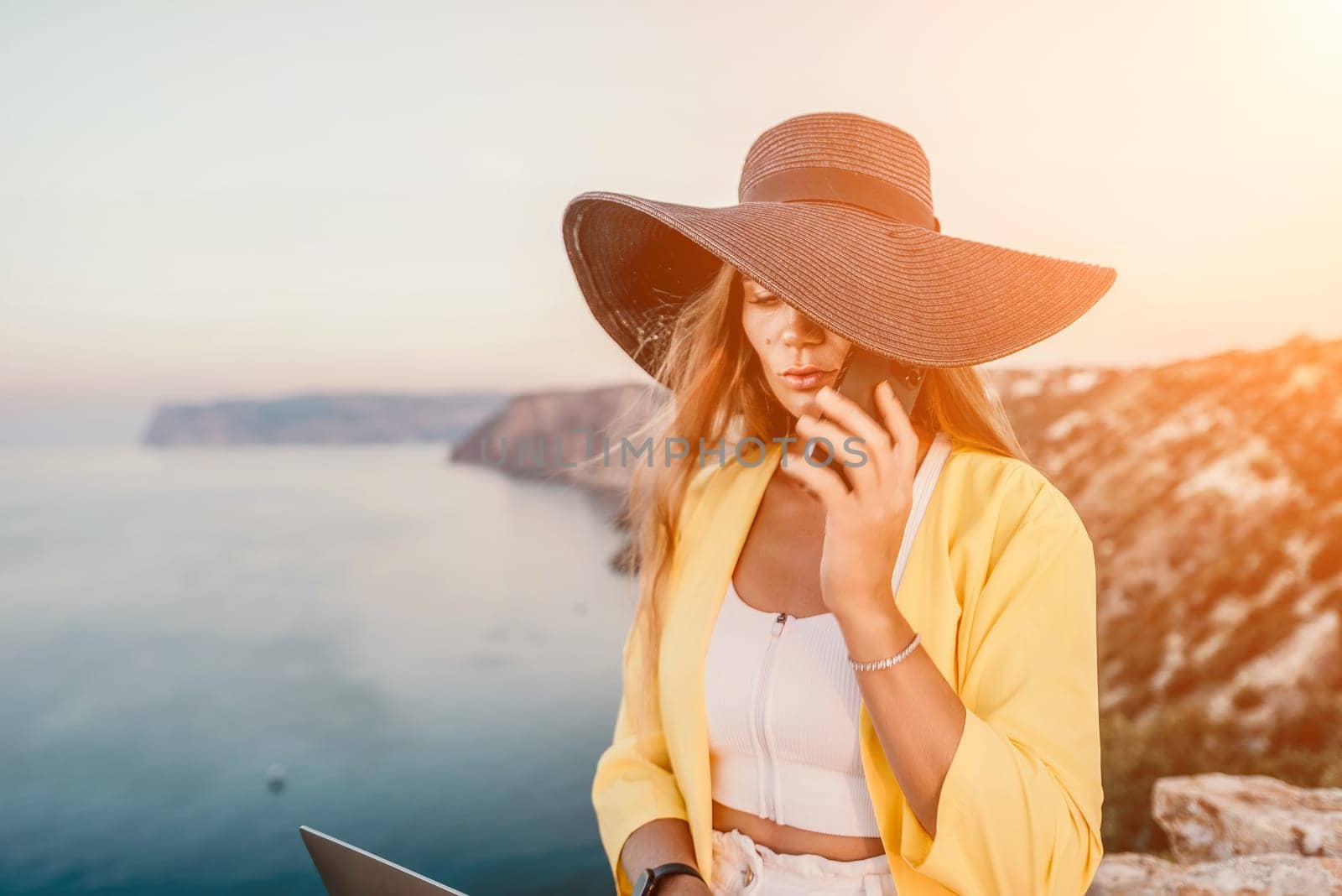 Successful business woman in yellow hat working on laptop by the sea. Pretty lady typing on computer at summer day outdoors. Freelance, travel and holidays concept.