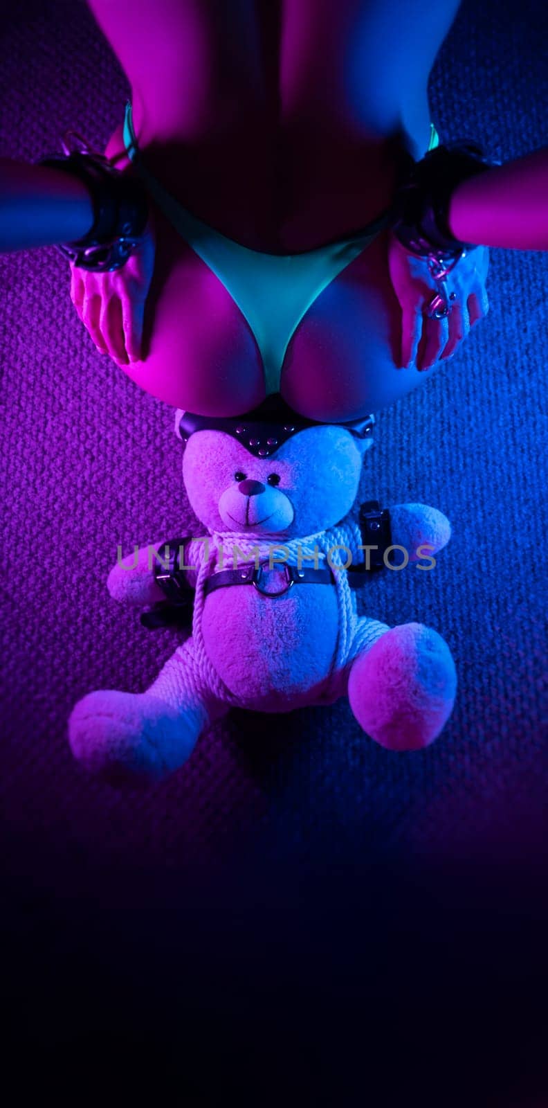 sexy buttocks of a girl in panties and hands behind her back in handcuffs , in an adult BDSM sex game and a teddy bear toy in the neon light
