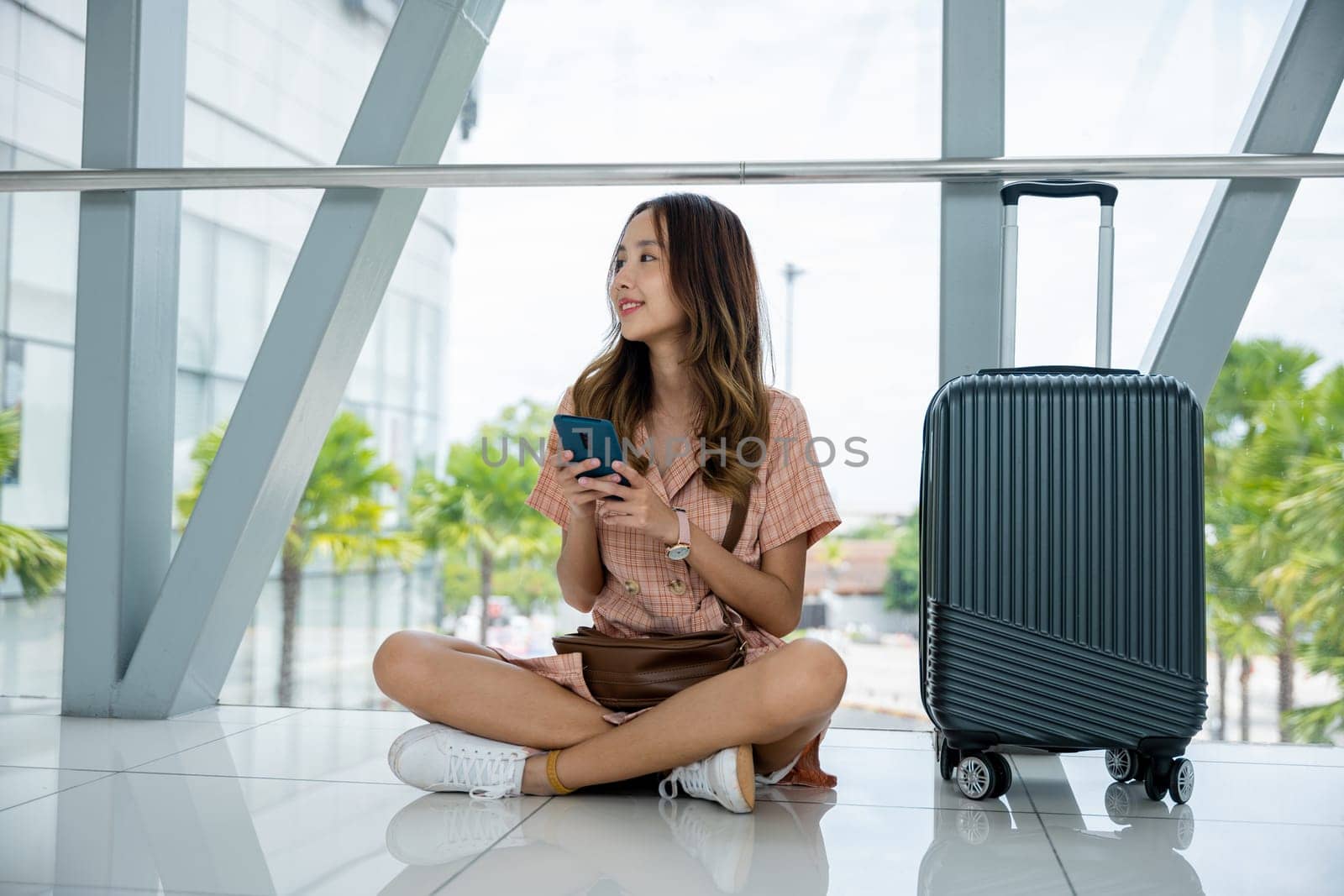 Asian woman sitting on the floor with her luggage and using her phone to stay connected while traveling. Tourist journey trip concept