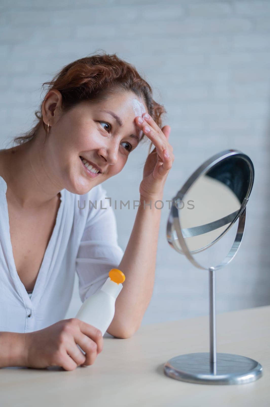 Portrait of a Caucasian woman with vitiligo uses sunscreen. A girl with a white pigment spot on her forehead looks in the mirror and is smeared with cream