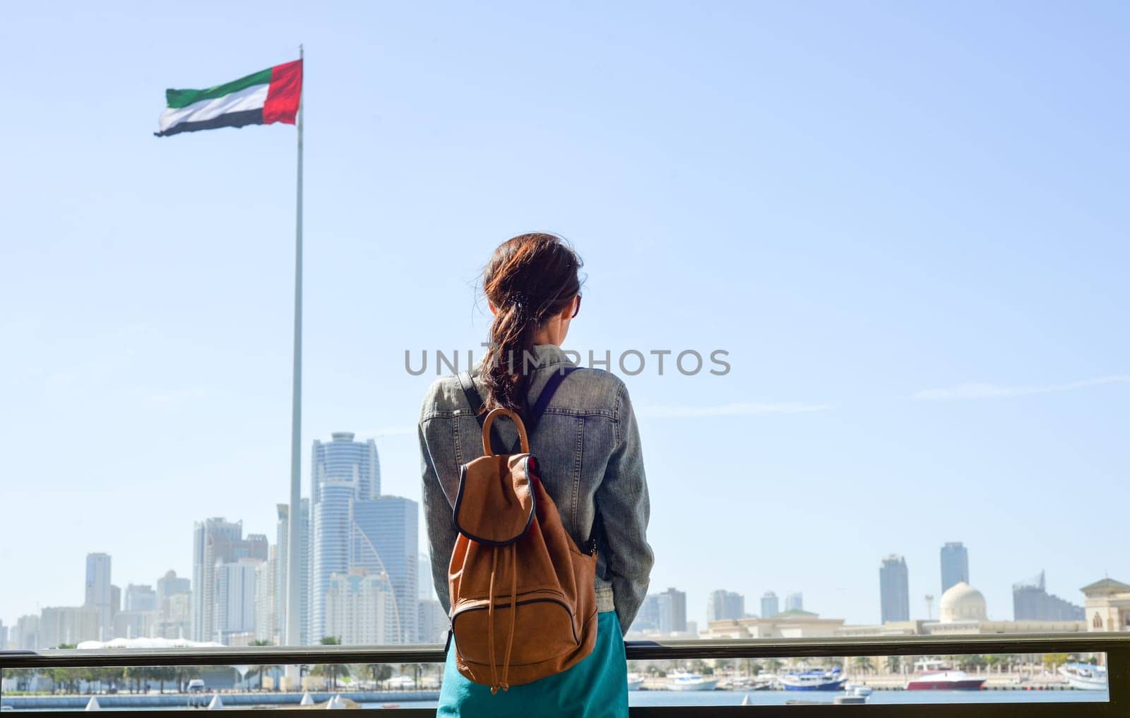 A tourist woman with a backpack admires the cityscape of Flag Island, Sharjah. Rear view of a woman on the embankment and the UAE flag. by Ekaterina34