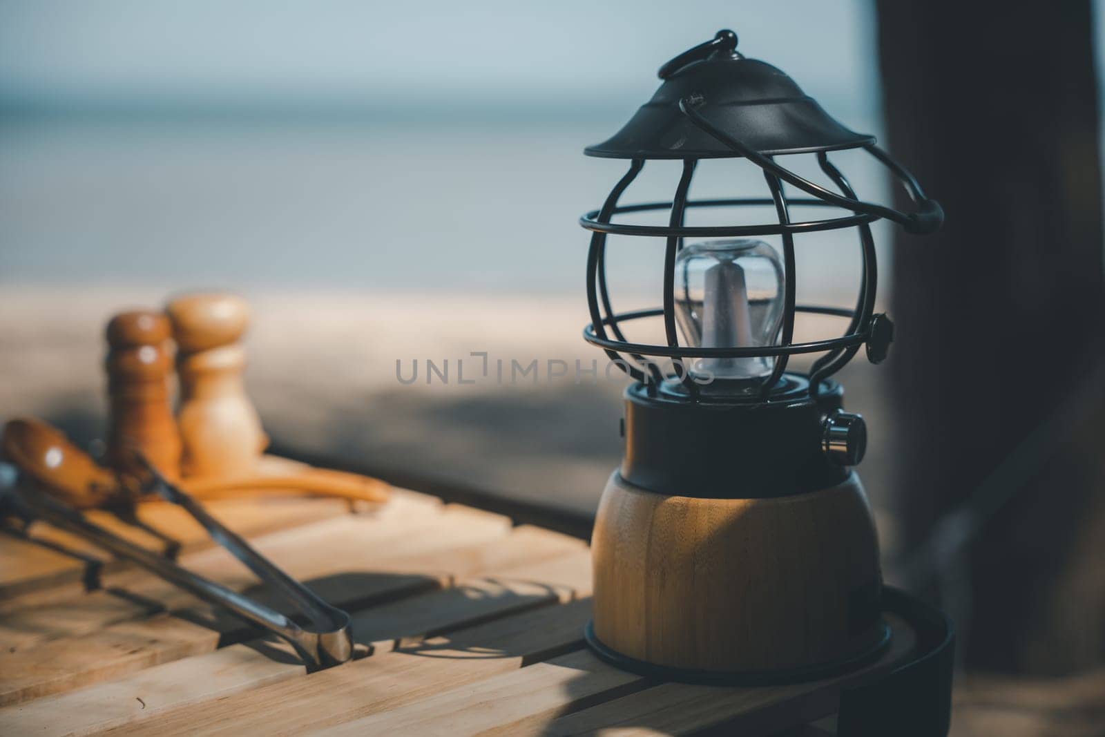 Camping by the beach with antique charm, A kerosene lamp and a modern LED lantern on a wooden table. Nature's beauty and modern equipment make for a unique experience.