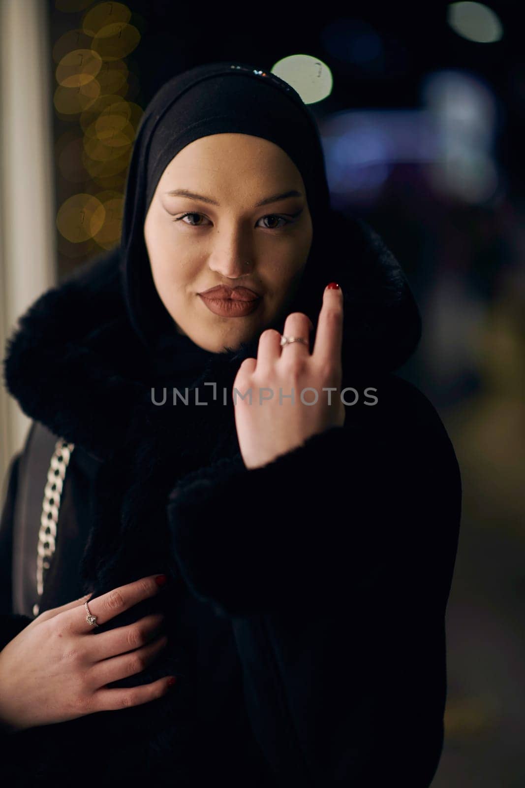 Young Muslim woman walking on urban city street on a cold winter night wearing hijab scarf veil a fashionable coat with bokeh city light in the background.