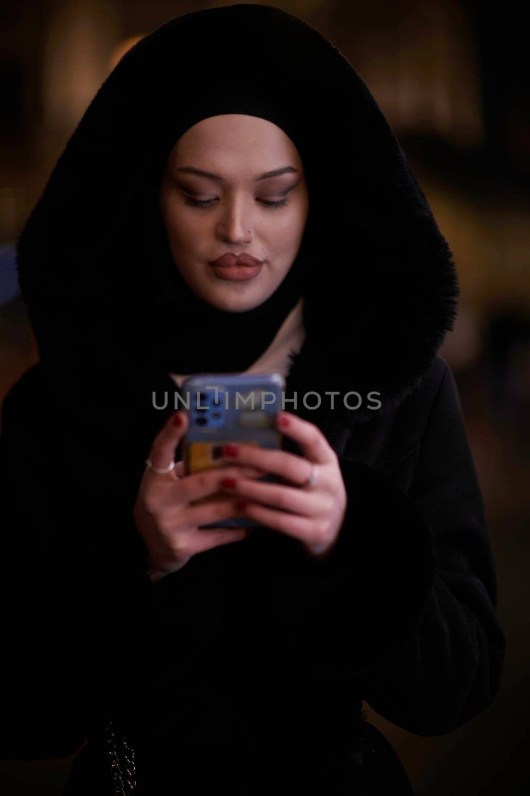 Young Muslim woman walking on urban city street on a cold winter night wearing hijab scarf veil a fashionable coat with bokeh city light in the background.