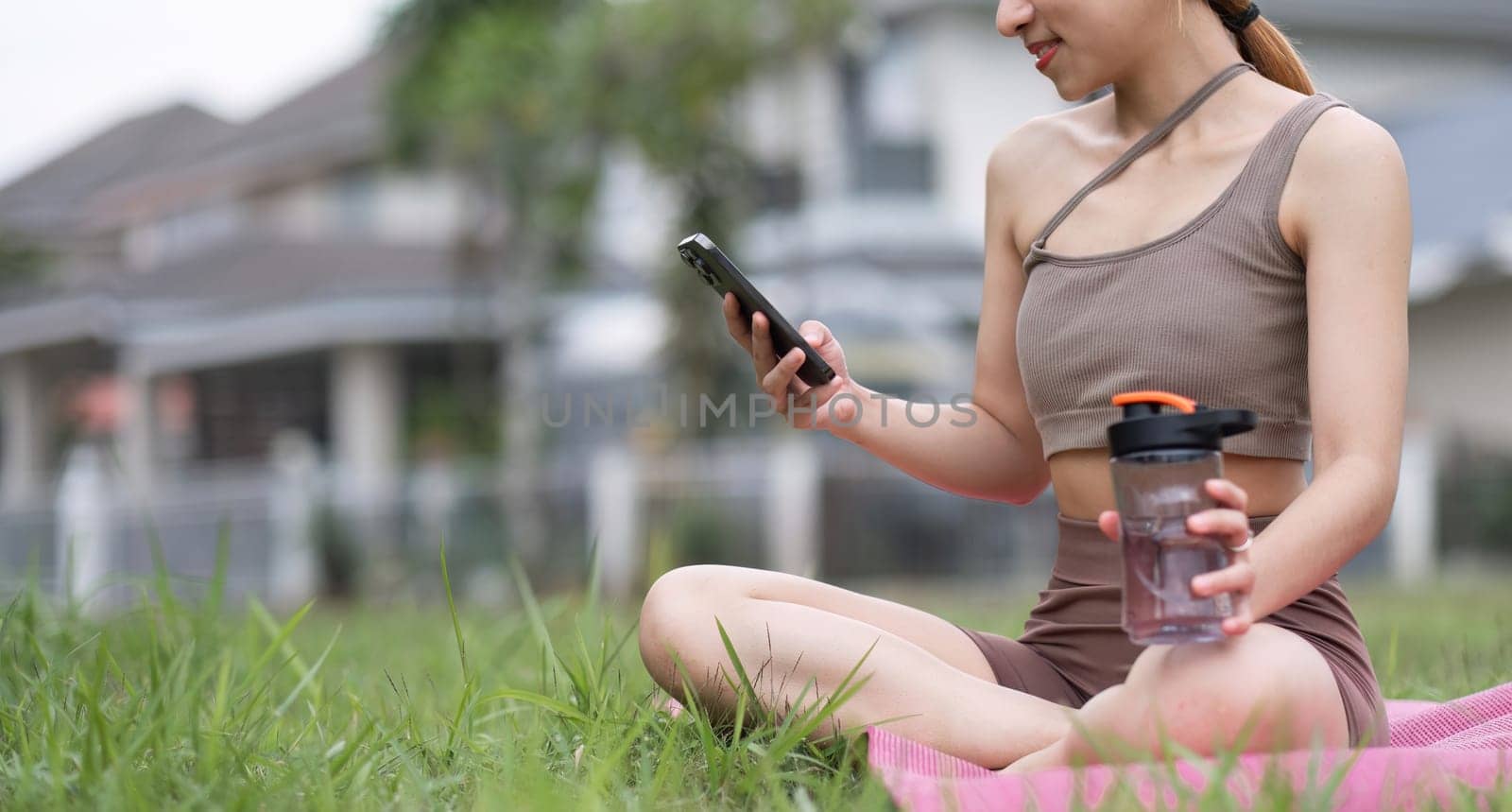 Young woman sitting on an exercise mat Use a smartphone during an outdoor yoga activity on the grass at a park. by wichayada